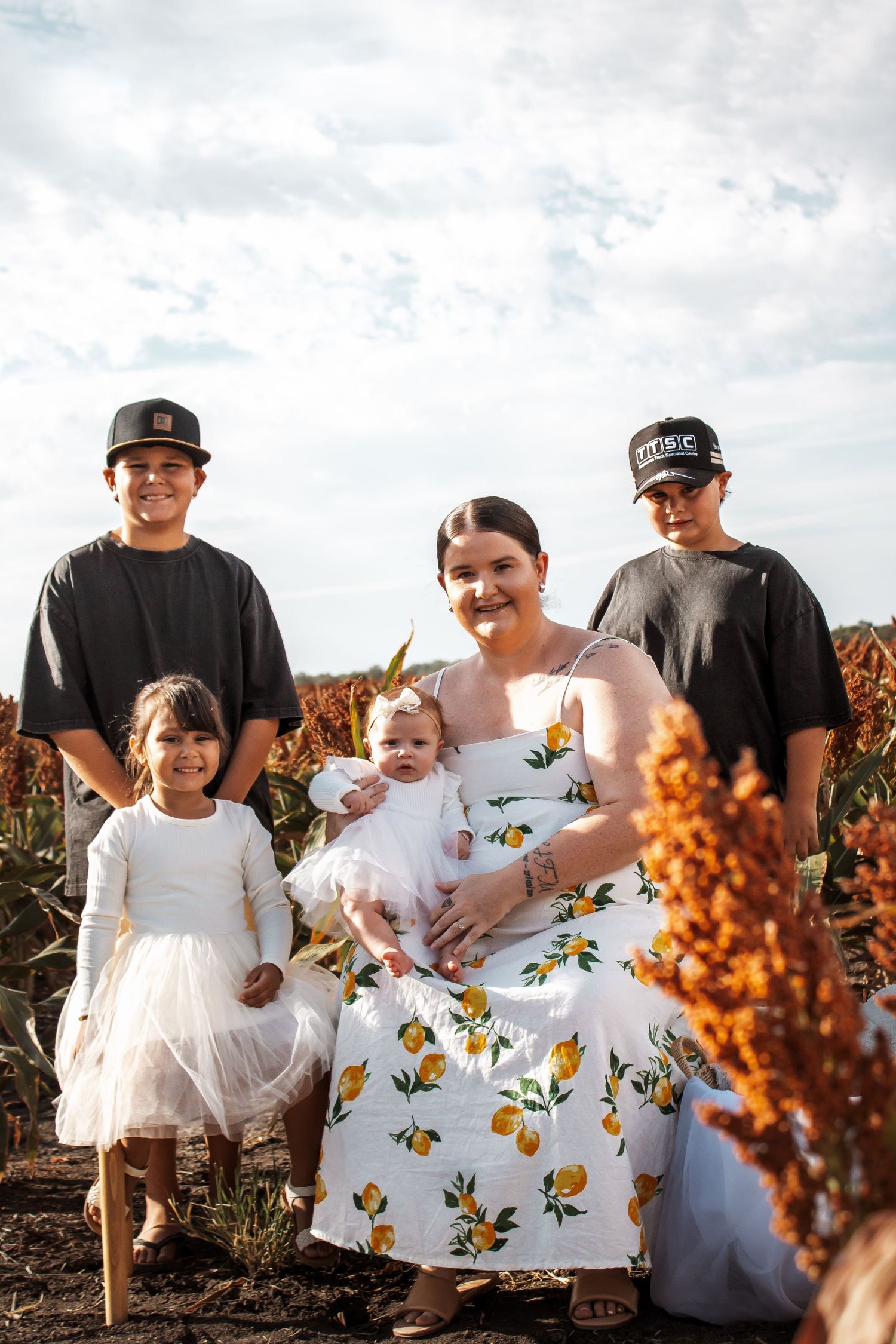 Family wearing coordinated white outfits sits together in a field with orange sorghum plants at sunset.