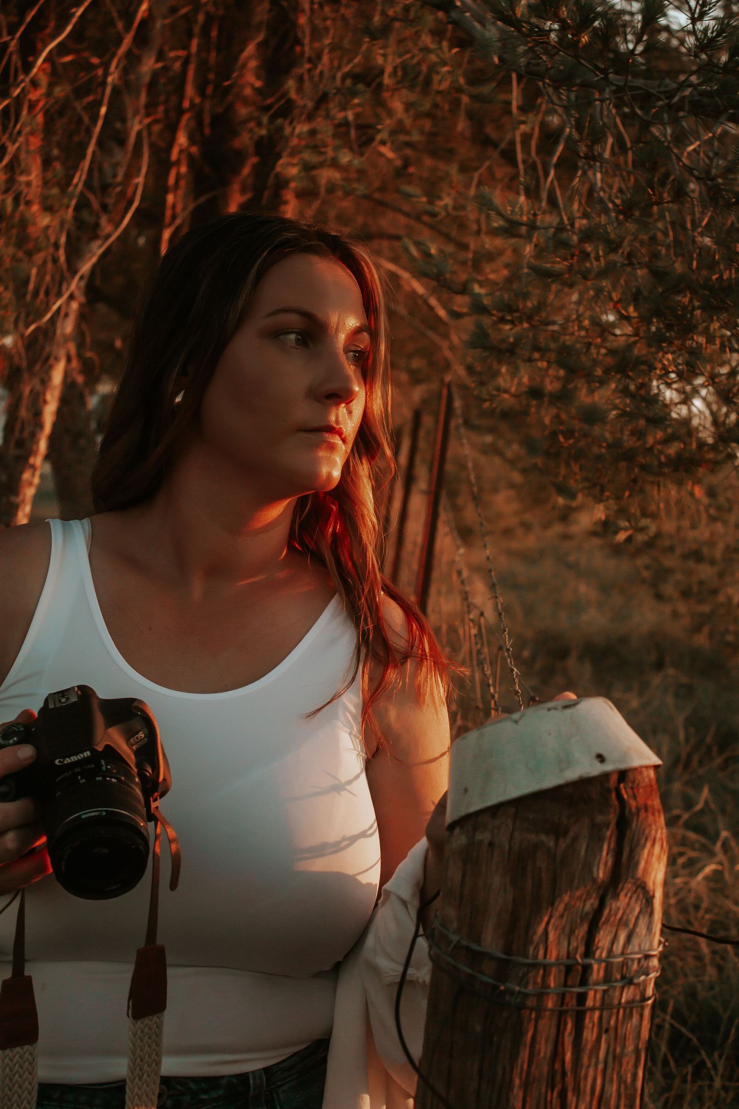 A person in a white tank top poses in warm sunset lighting among trees and foliage.