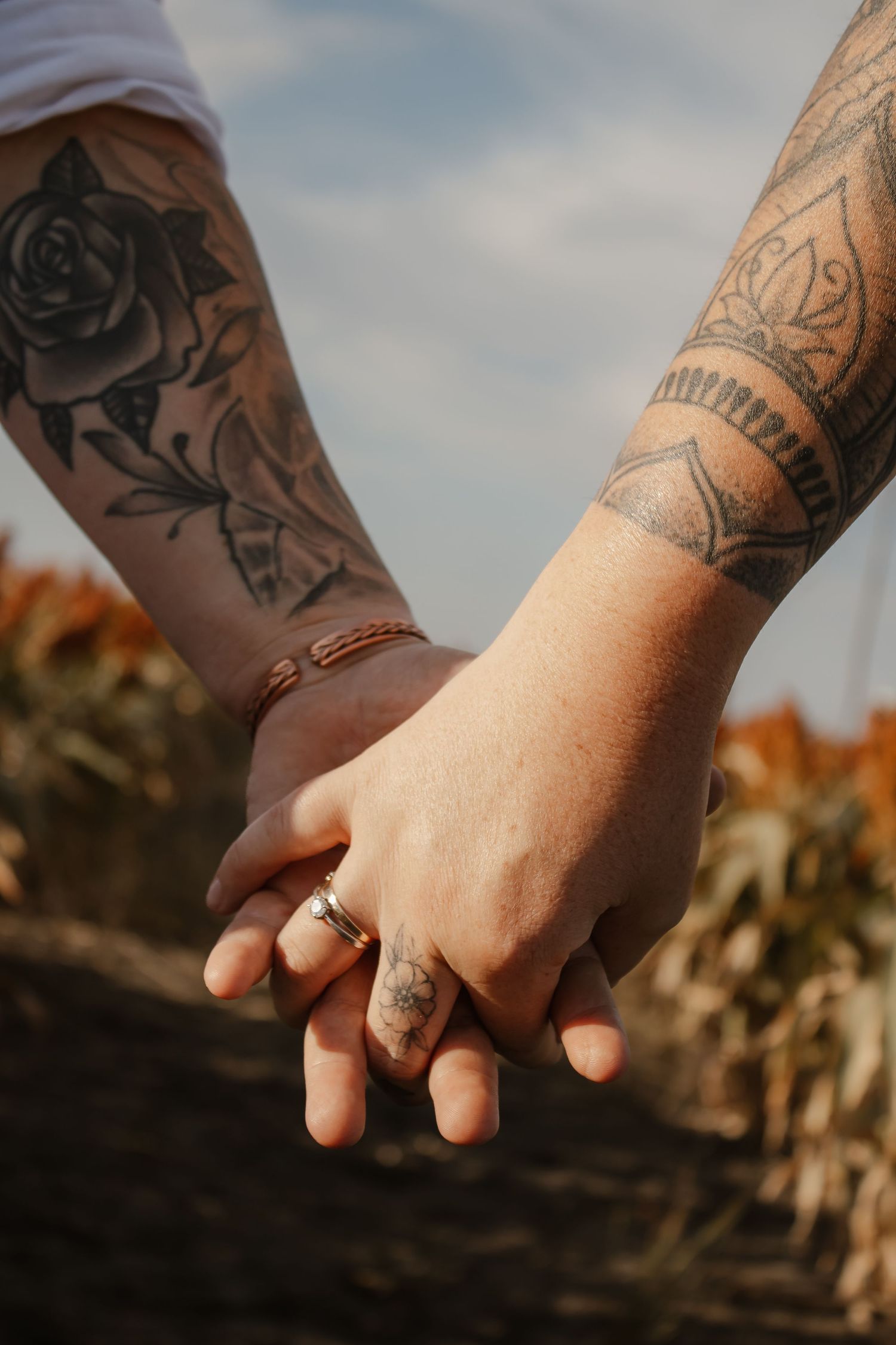 Close-up of two tattooed hands holding each other with wedding rings against a blurred outdoor background.