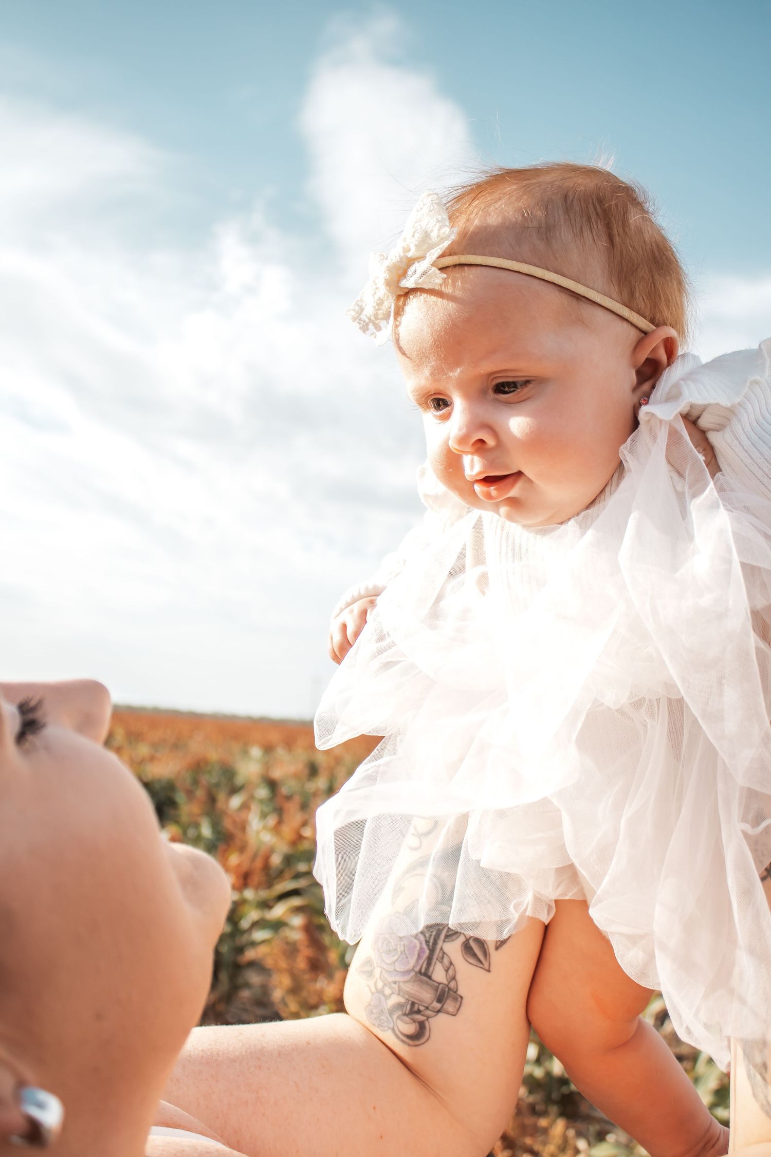 A baby wearing a white ruffled dress gazes upward against a bright blue sky with fluffy clouds.