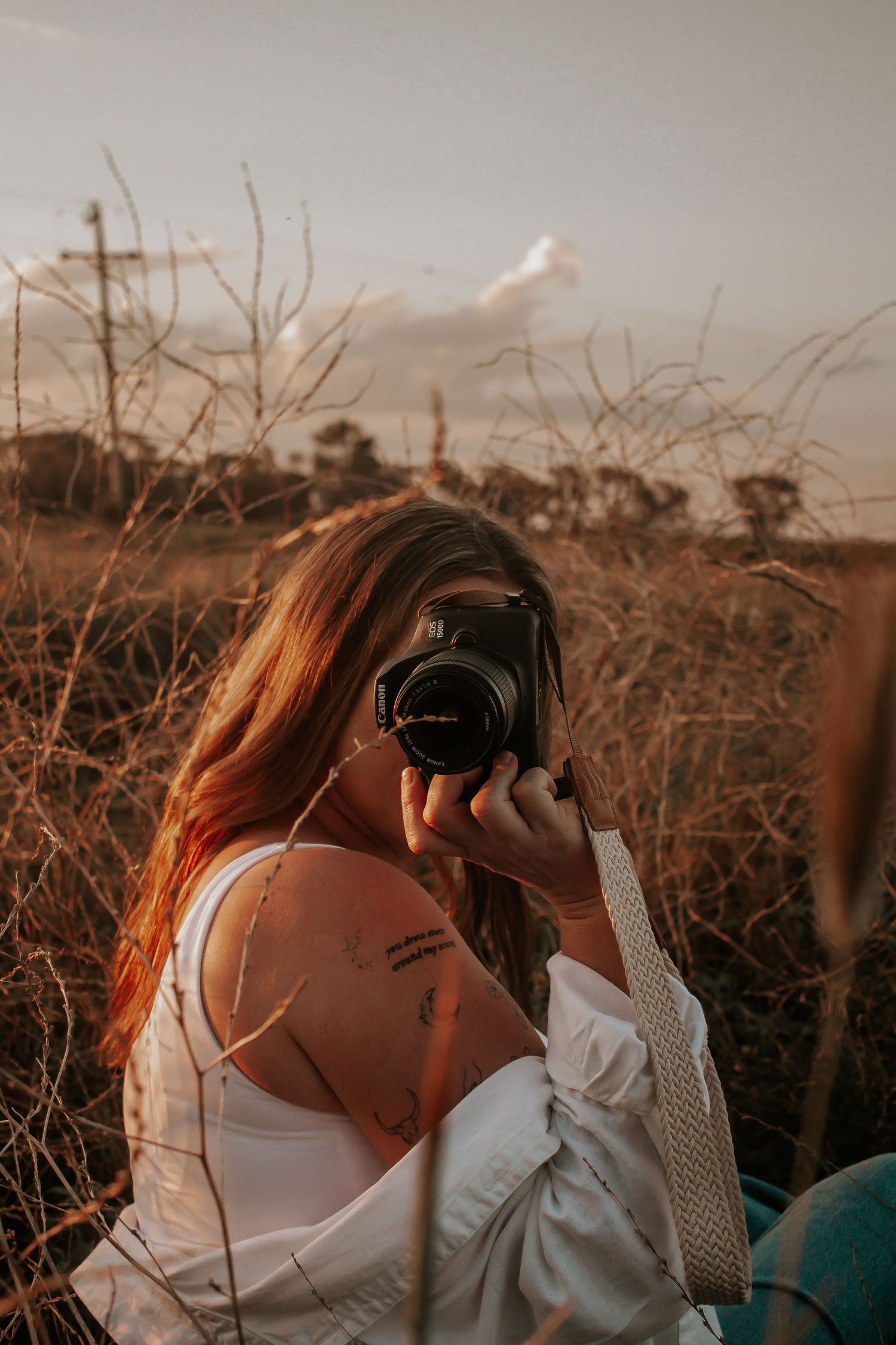 Photographer in white top taking photos during golden hour against bare branches in sepia-toned lighting.