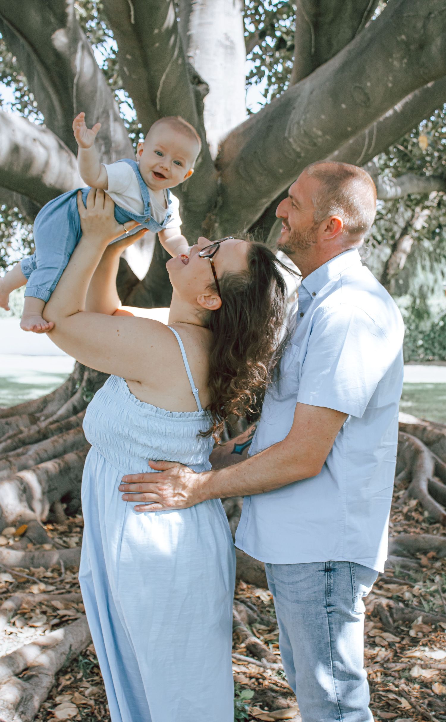 A couple shares tender moments with their toddler under a large tree in a series of candid outdoor family photos.