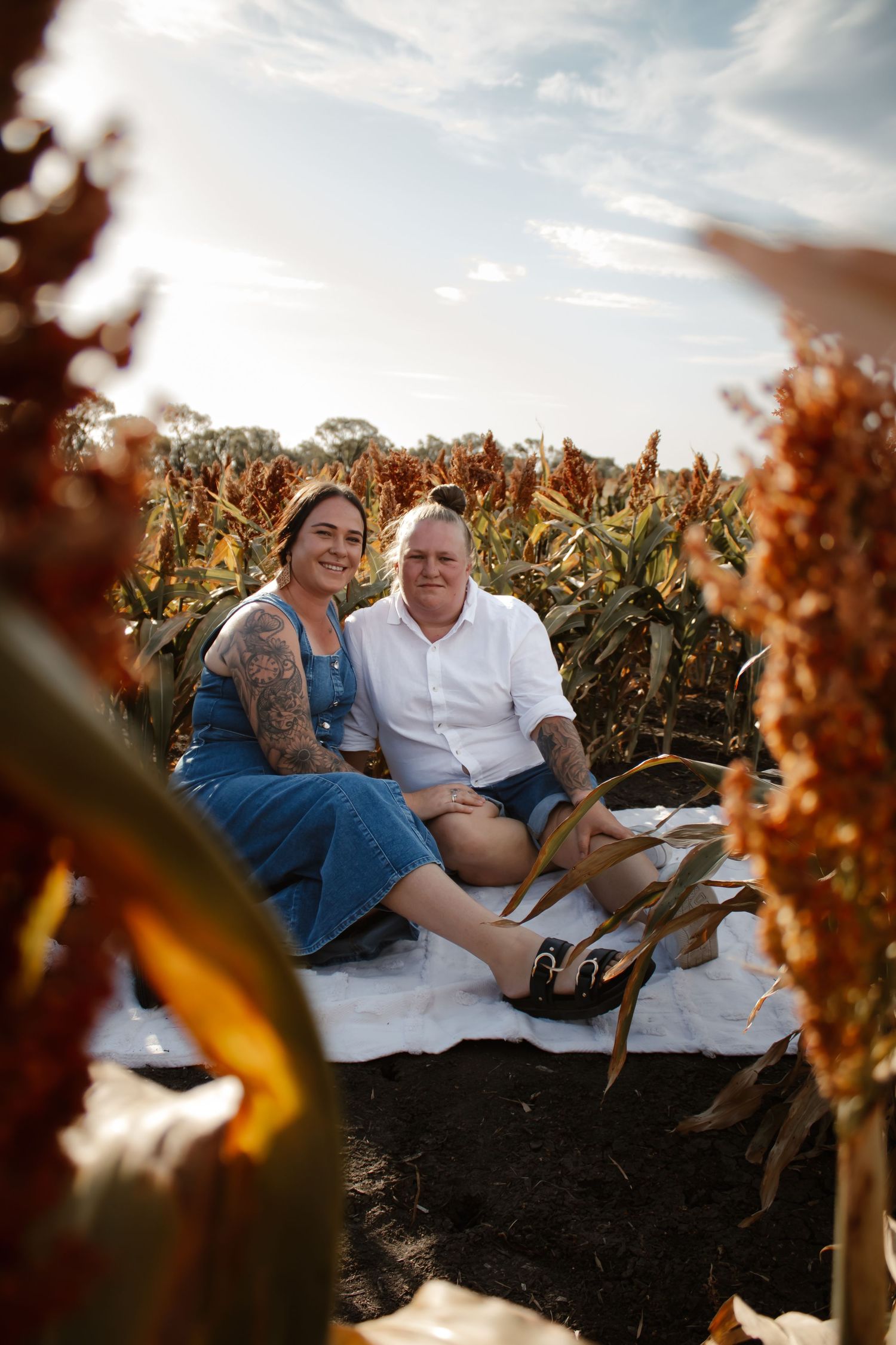 A couple sits together on a blanket in a field during golden hour with autumn foliage in the background.