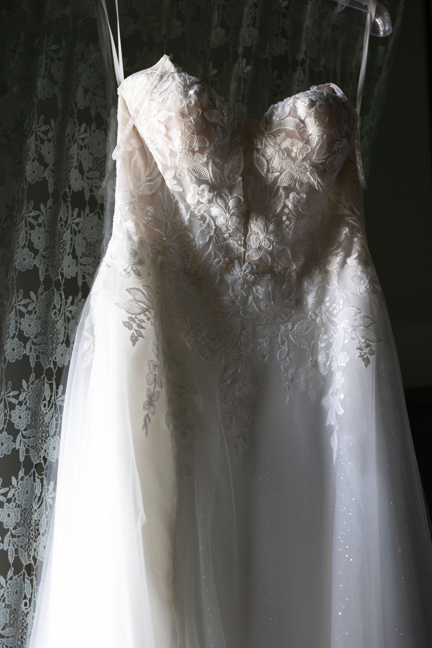 Elegant white lace wedding dress with intricate floral detailing hangs on display against a dark background.