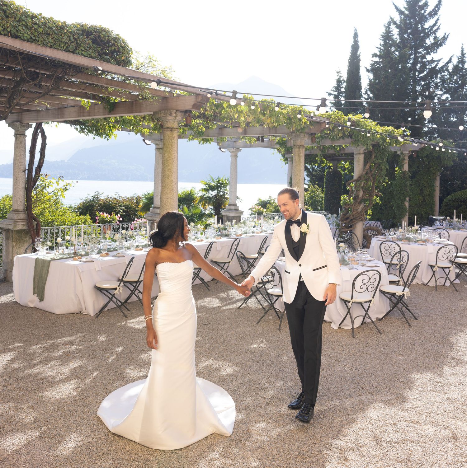 A panoramic view of an outdoor wedding venue with white chairs arranged under a rustic pergola by the sea.