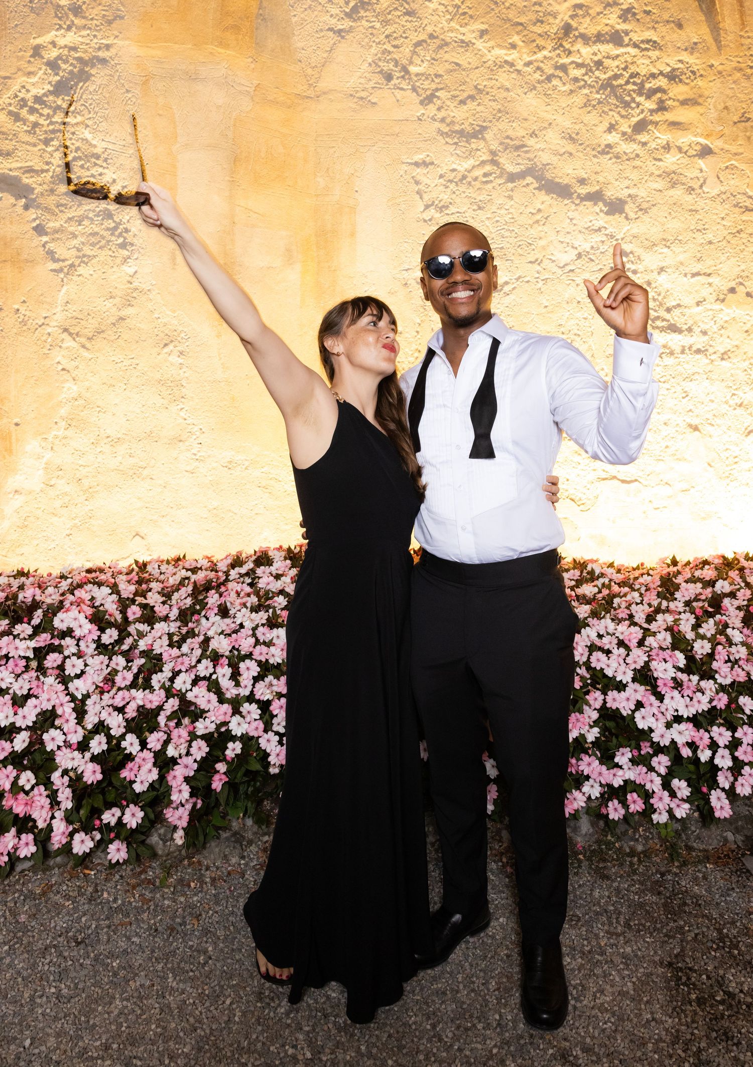 Two people in formal attire dancing and posing against a yellow wall with pink flowers at the bottom.
