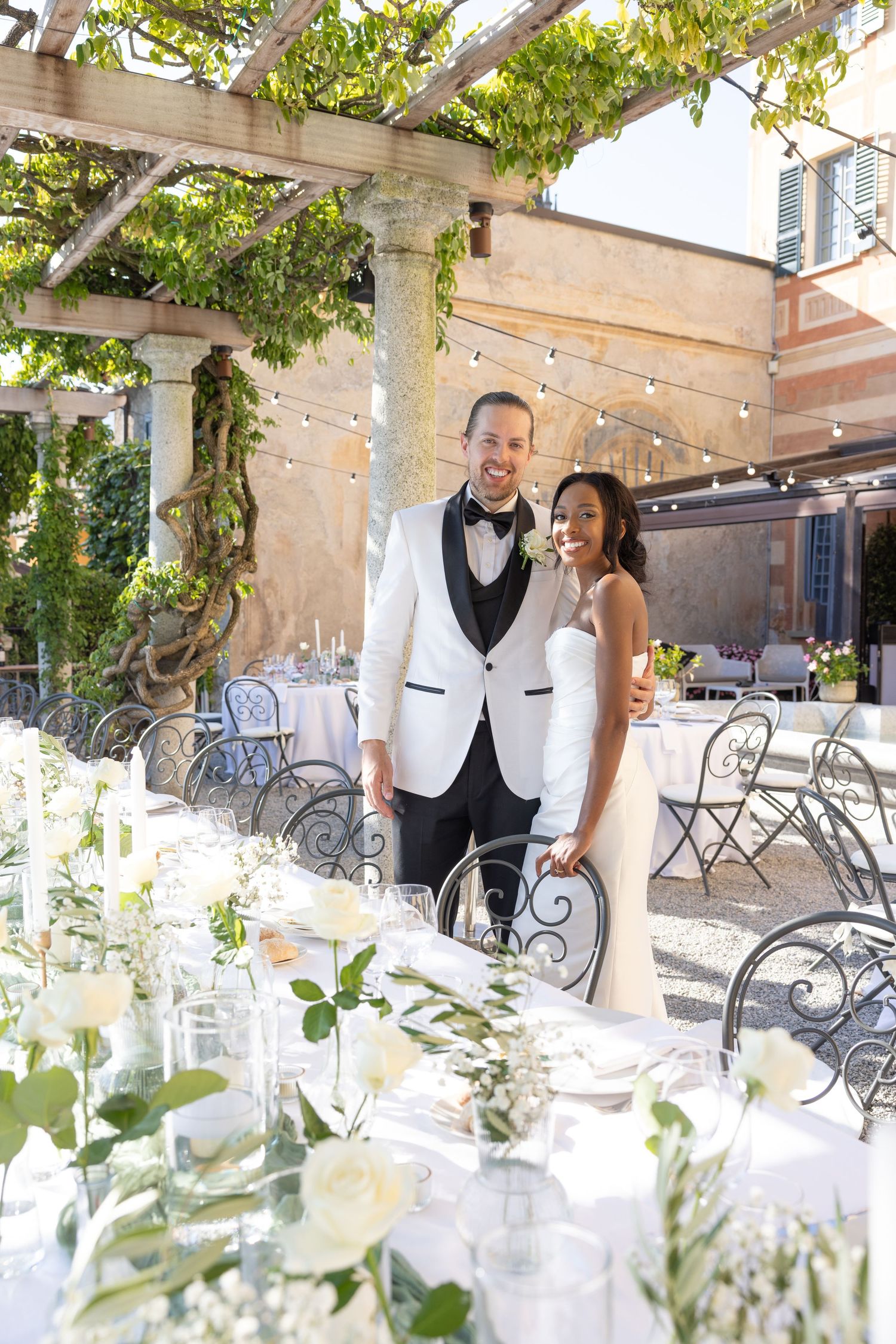 Elegant outdoor wedding reception setup with white floral arrangements and string lights under a vine-covered pergola.