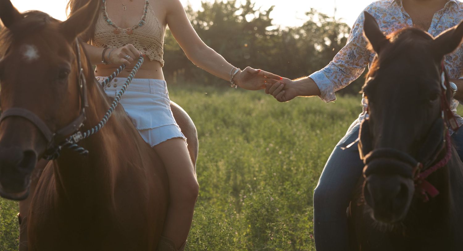 Two people riding horses side by side reach for each other's hands in golden evening light.