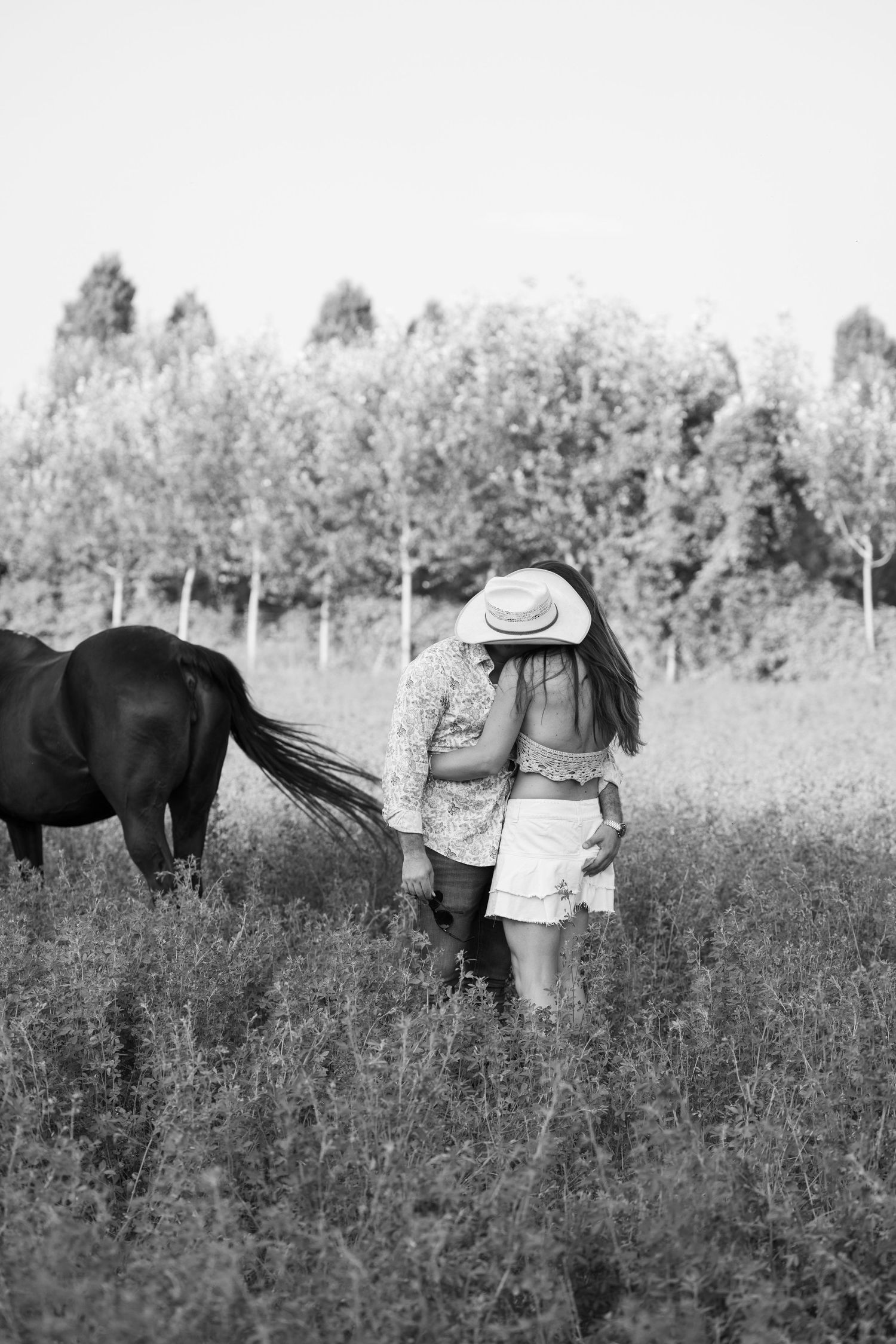 Black and white photo of couple with horses in a wildflower meadow wearing western attire.