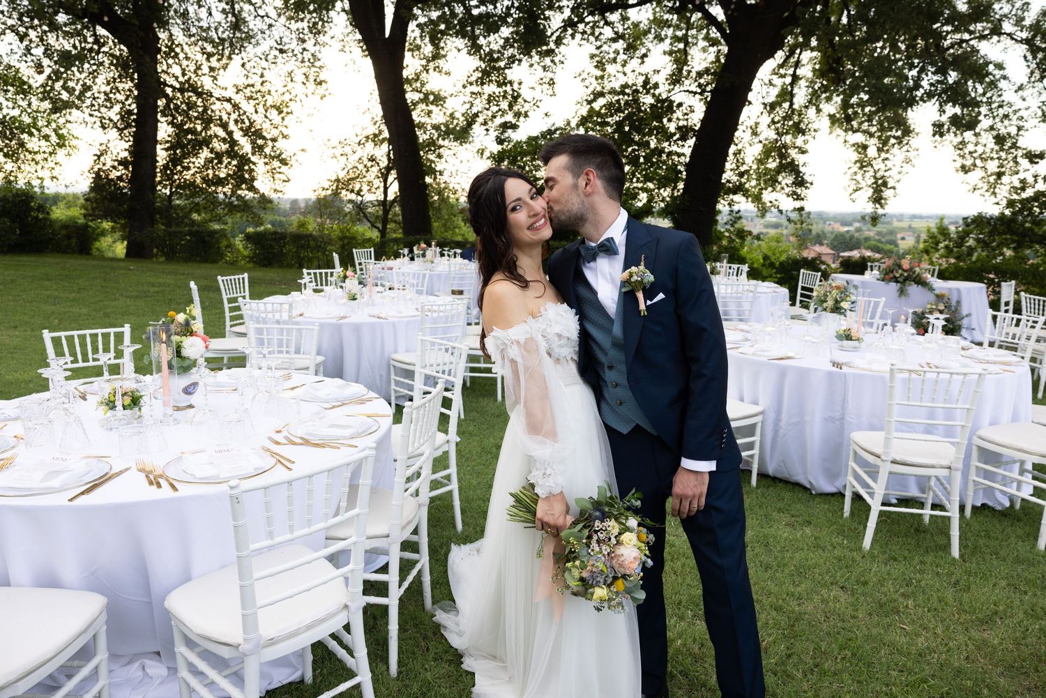Elegant outdoor wedding reception with white Chiavari chairs and table settings on a grassy lawn.