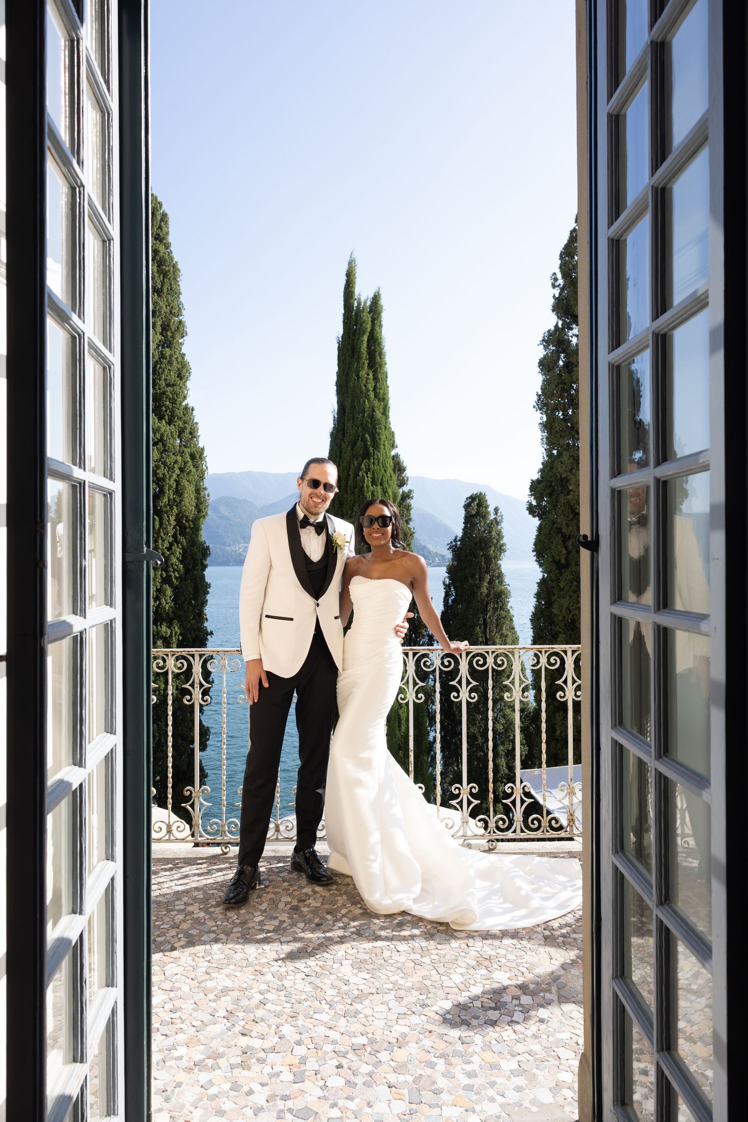 A romantic wedding scene through French doors showing a couple on an elegant balcony overlooking a lake and mountains.