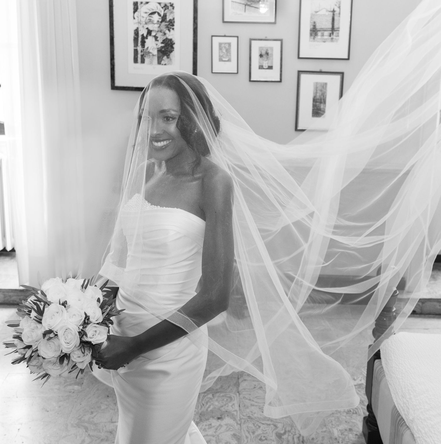 A bride in a white wedding dress adjusts her flowing veil while standing in a room with framed artwork on the walls.