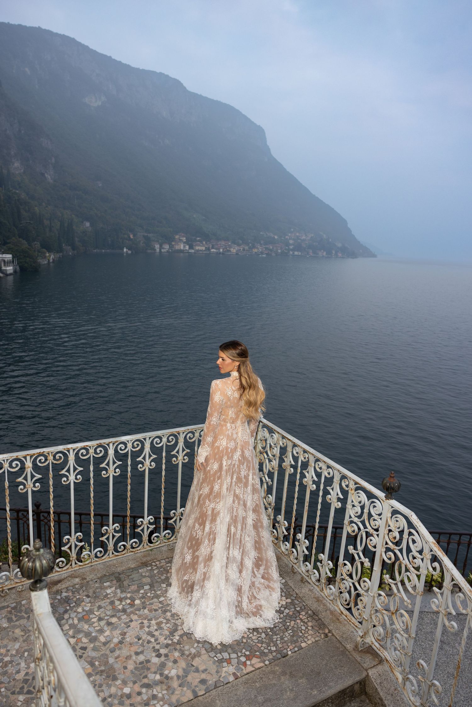 The Bride in a flowing dress stands on a stone balcony overlooking Lake Como with misty mountains in the background.