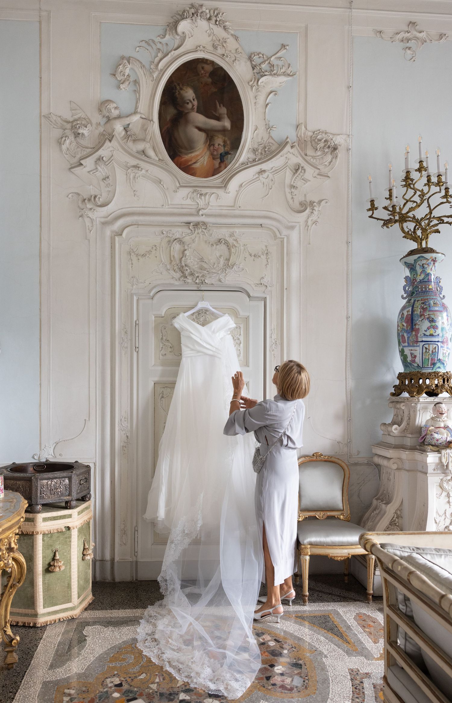 White wedding dress displayed in ornate baroque room with antique furnishings and decorative moldings.