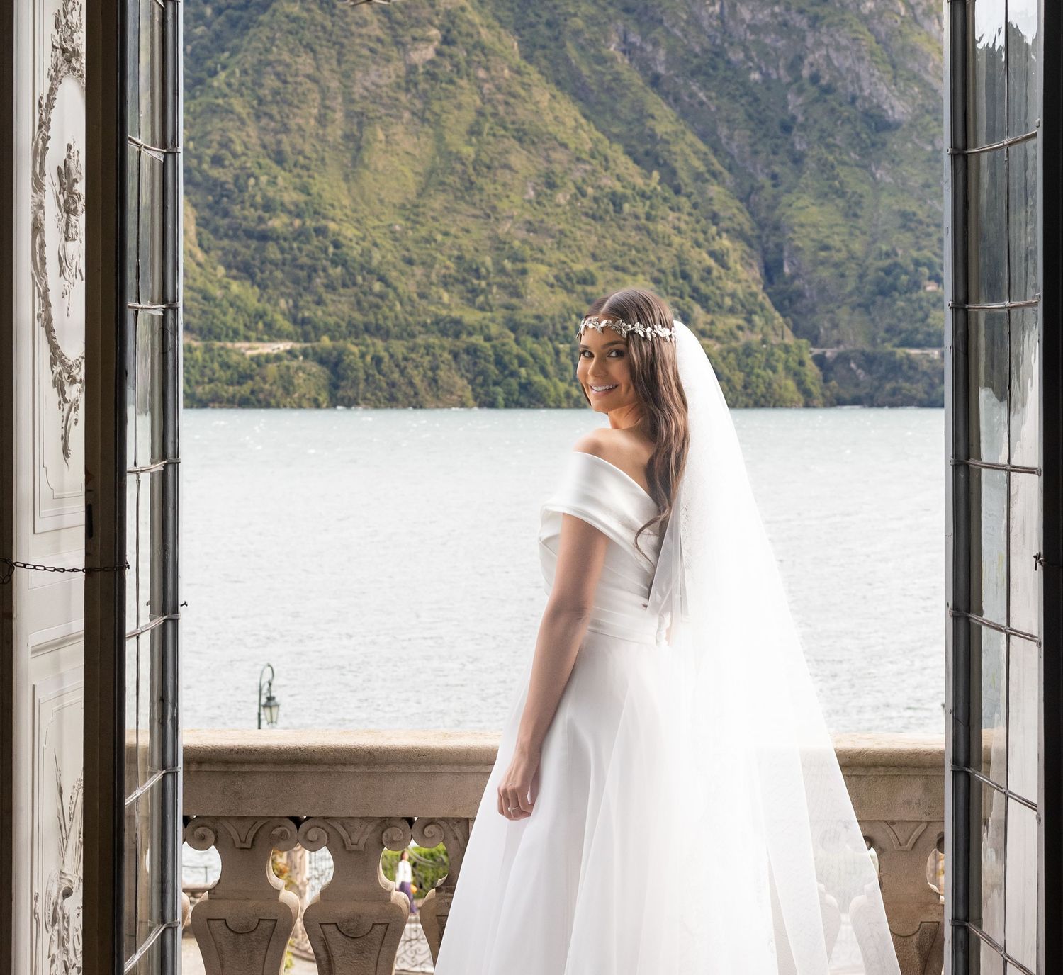 Wedding dress silhouetted against mountain lake view through grand balcony doors.