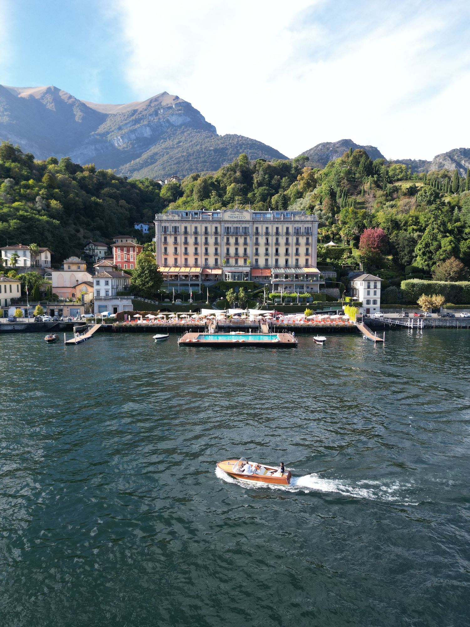 Luxury hotel Tremezzo and ferry dock on Lake Como with mountains in background and small boat cruising on blue waters.