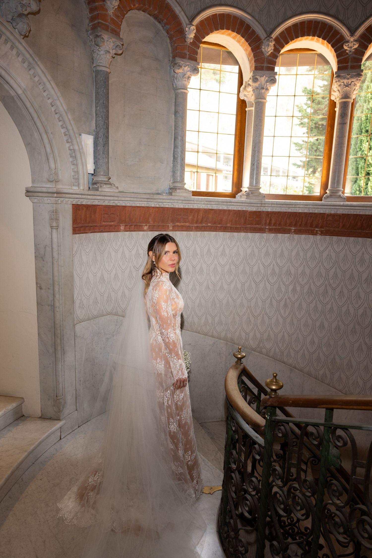 Elegant bridal photoshoot in a historic stone corridor with arched windows and ornate architectural details.