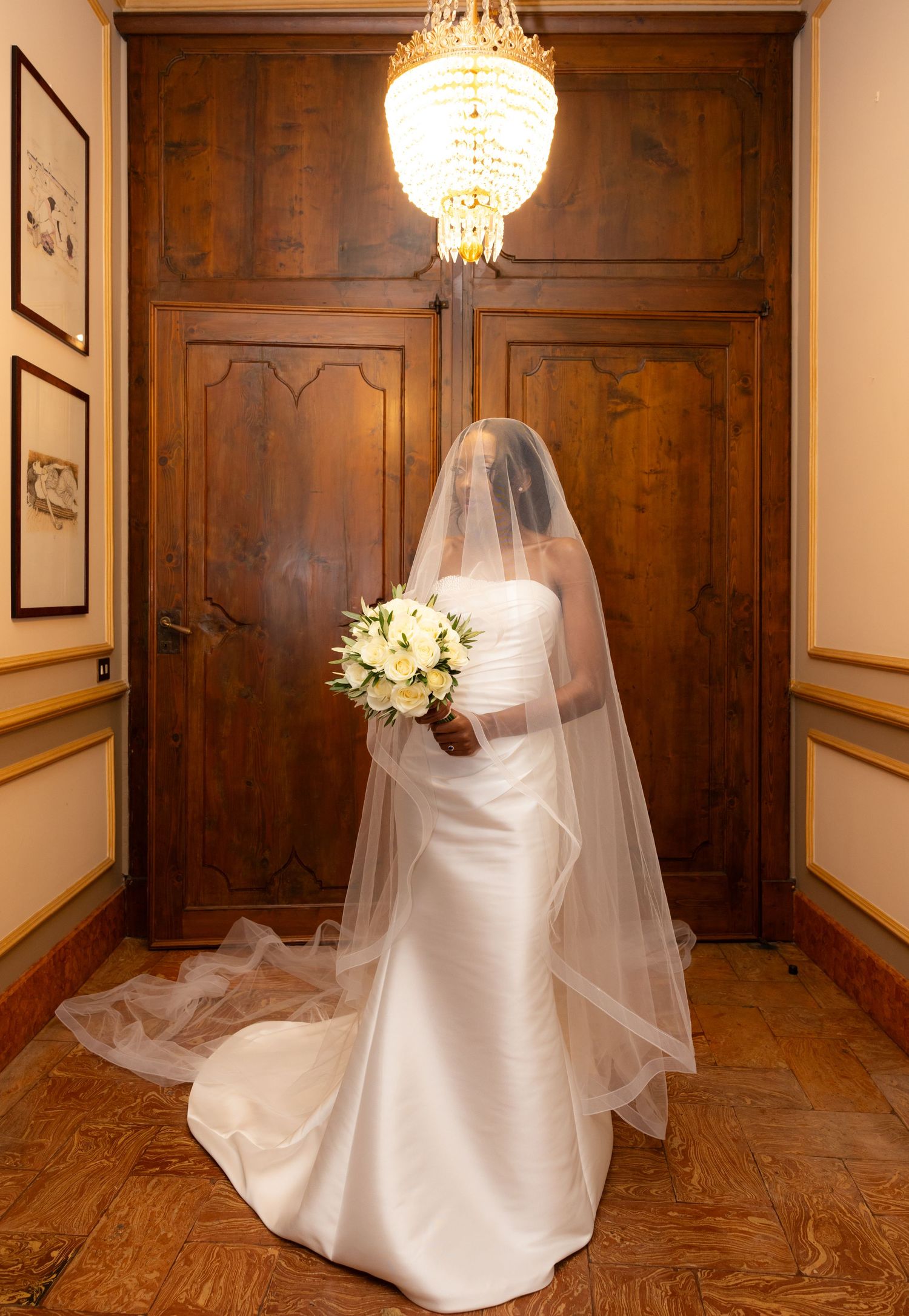 A bride in a flowing white wedding gown and veil poses in an elegant wood-paneled room with crystal chandeliers.