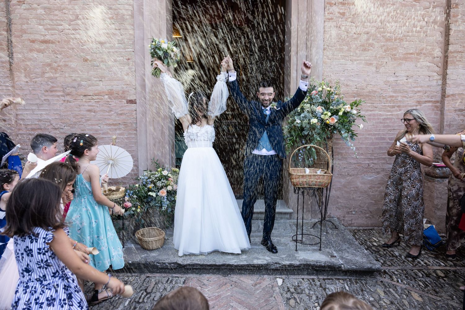 Wedding guests gather in a courtyard with brick walls and floral decorations for an outdoor celebration.