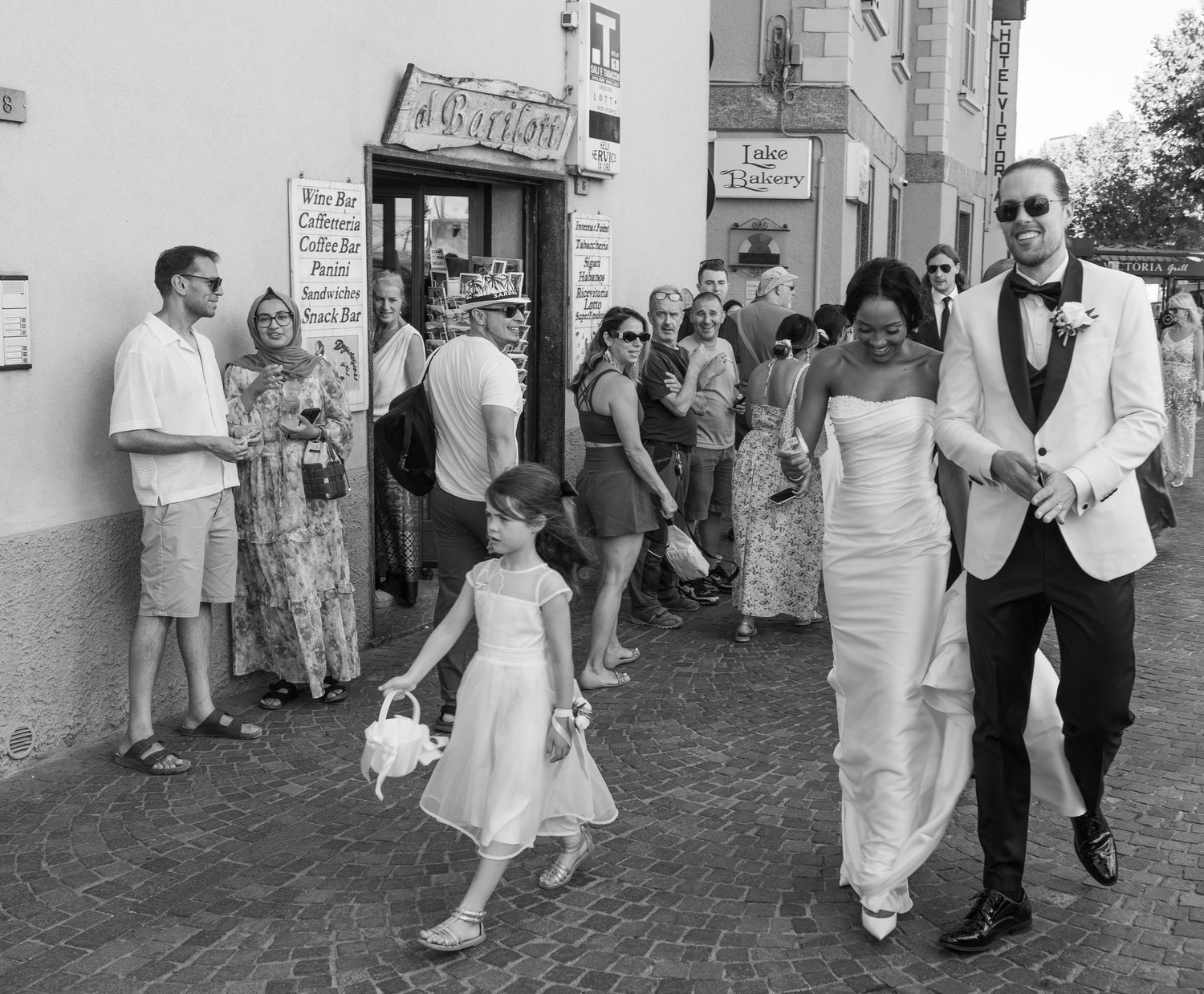 People in white dresses and formal attire walk along a brick sidewalk during a wedding celebration.