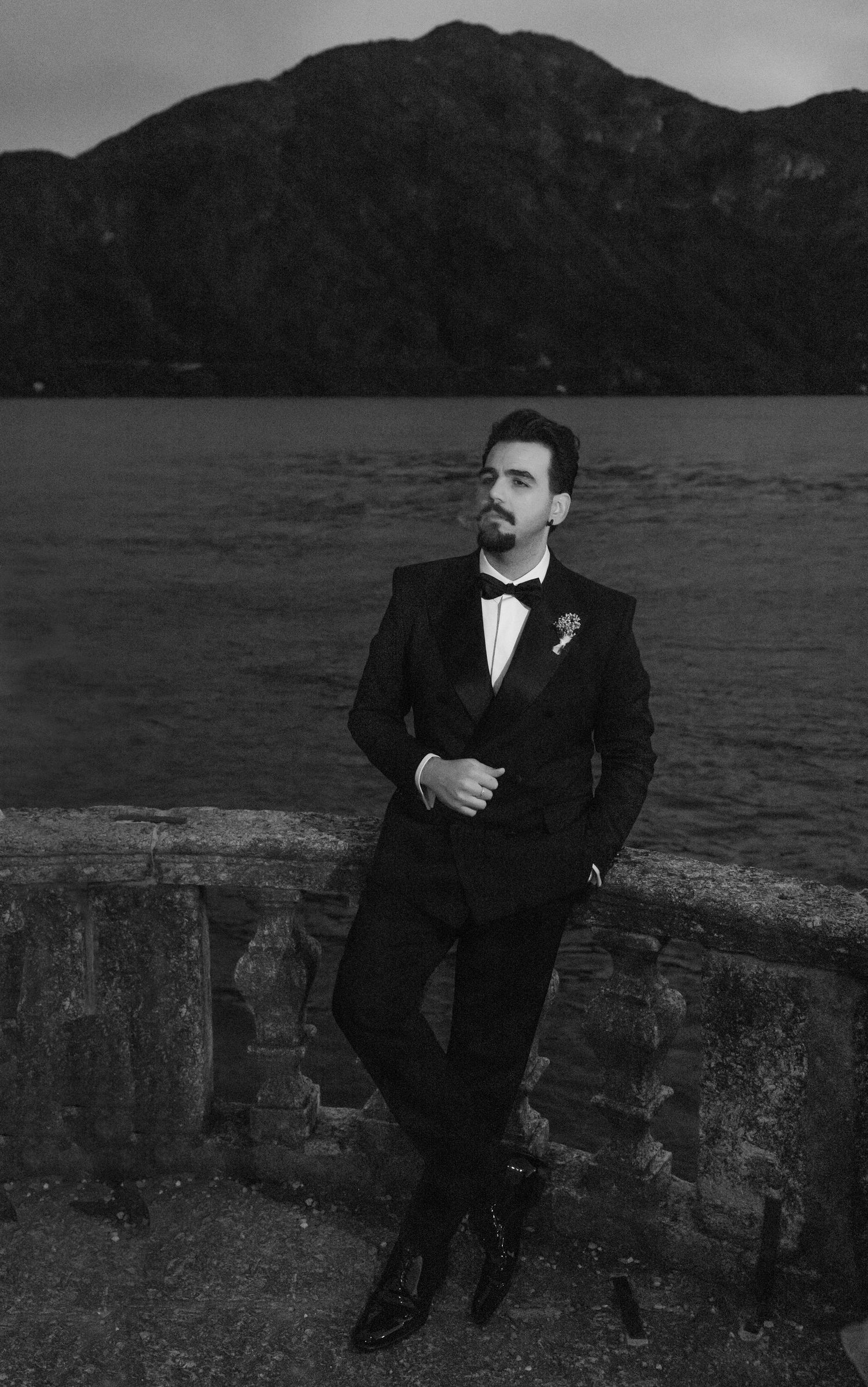 A black and white portrait against a stone wall with mountains and lake in the background.