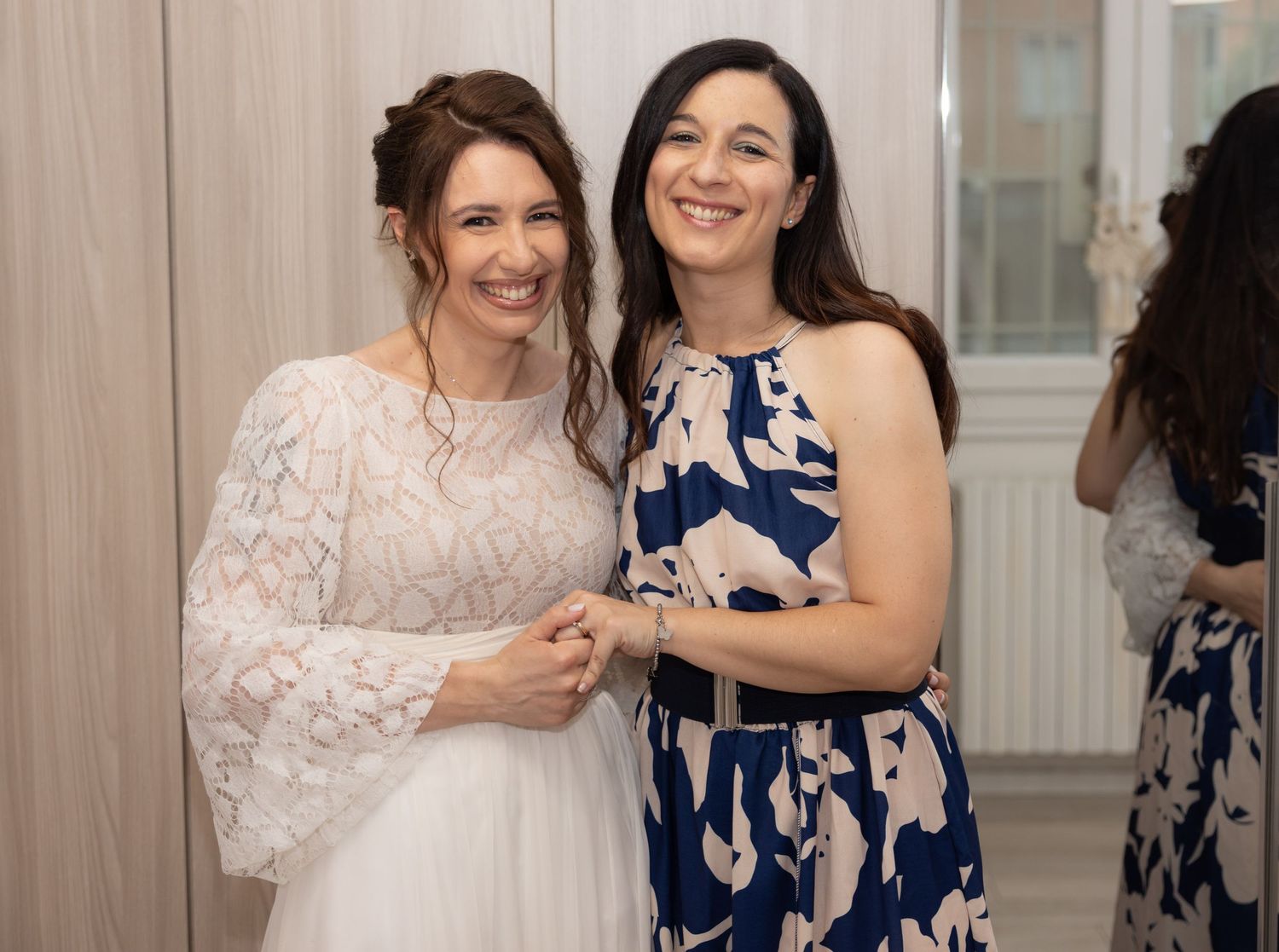 Two women share a joyful moment at a wedding, one wearing a white lace dress and the other in a blue floral dress.