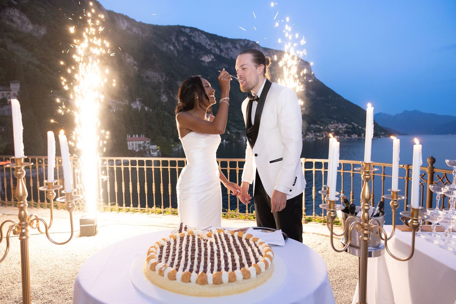 Panoramic view of sparklers illuminating an elegant outdoor wedding reception by the lake at dusk.