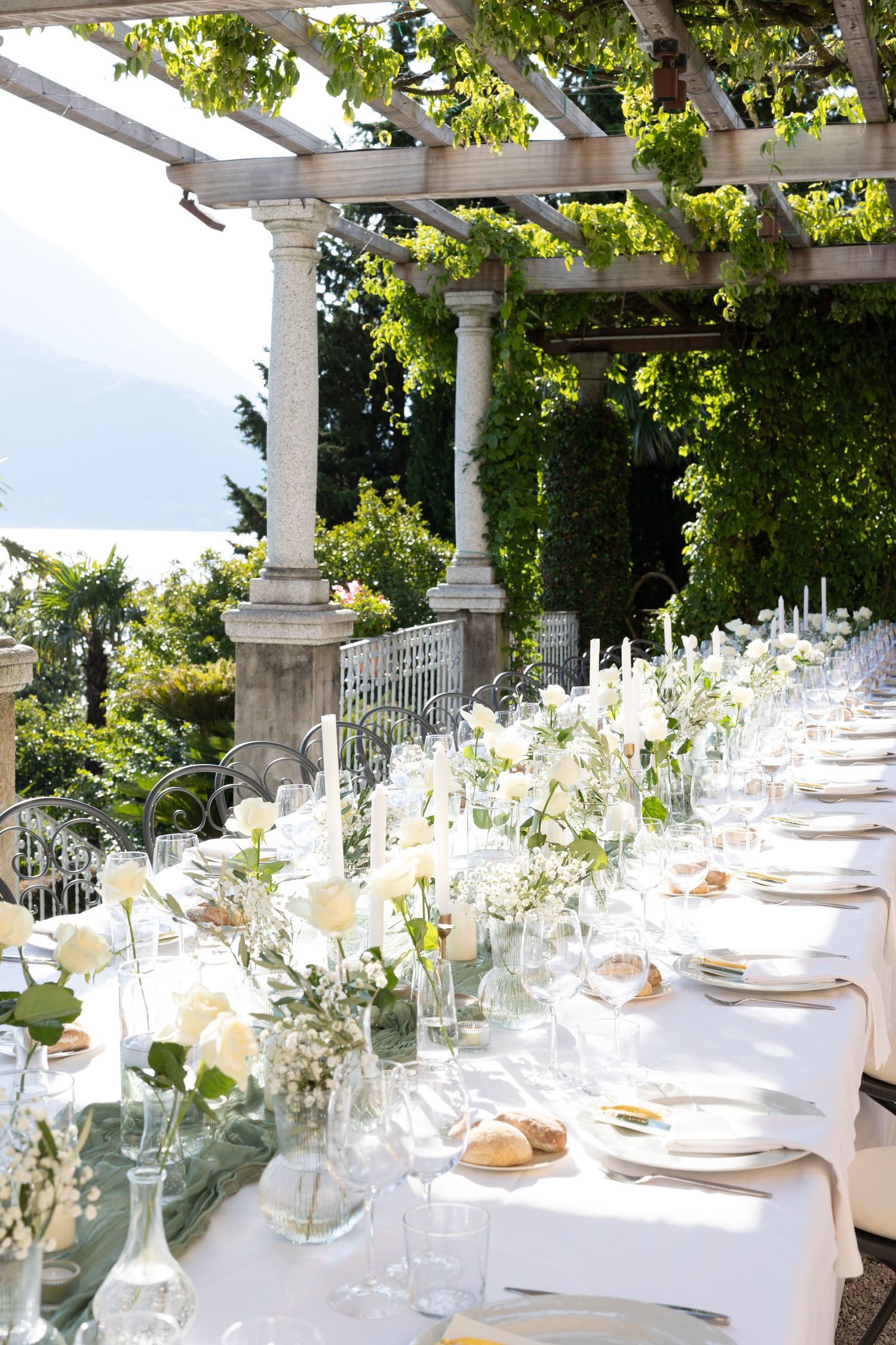 Elegant outdoor wedding reception table setup with white tablecloths and floral centerpieces under a pergola.