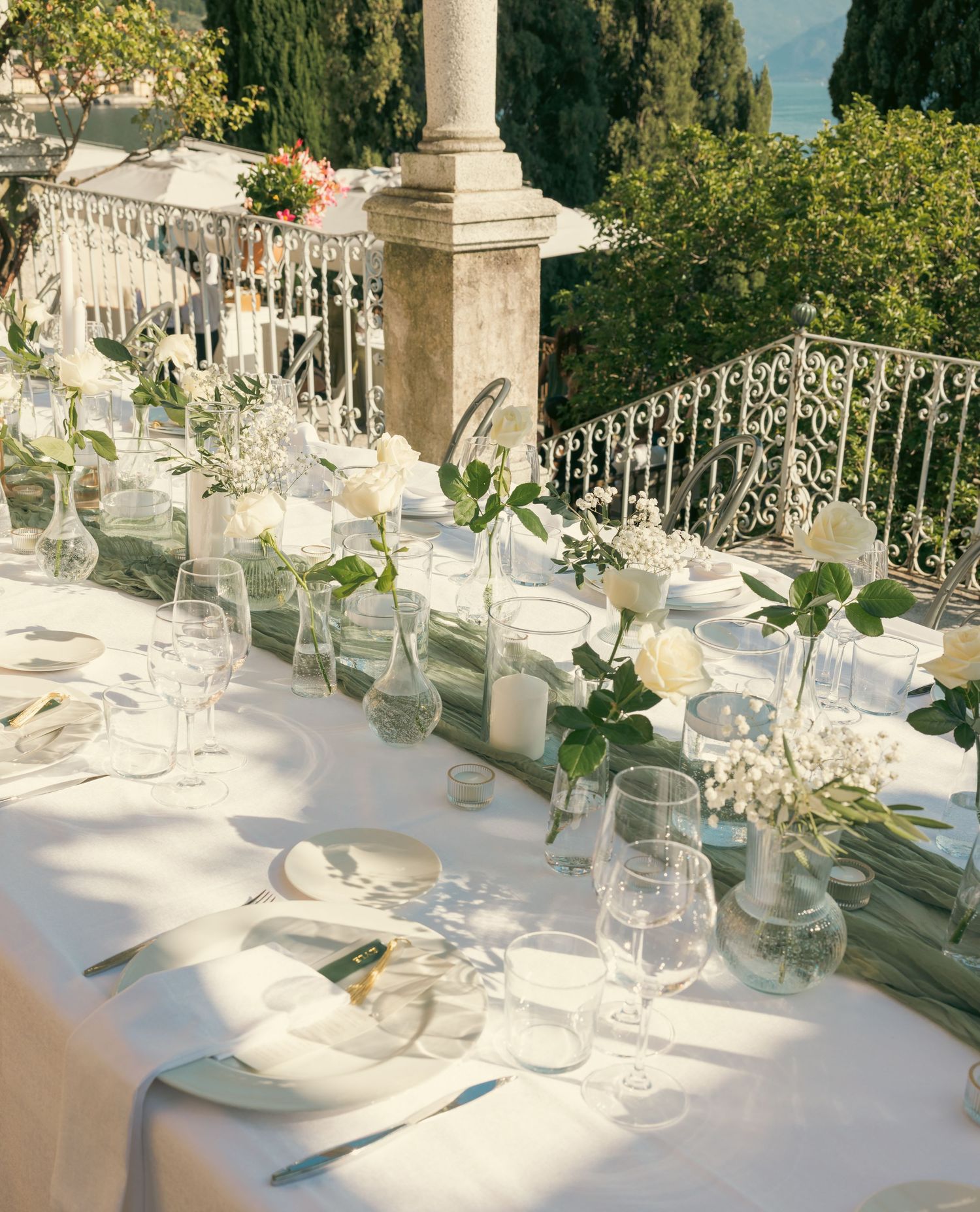 Elegant outdoor wedding table setting with white linens, glassware and floral arrangements along a stone balustrade.