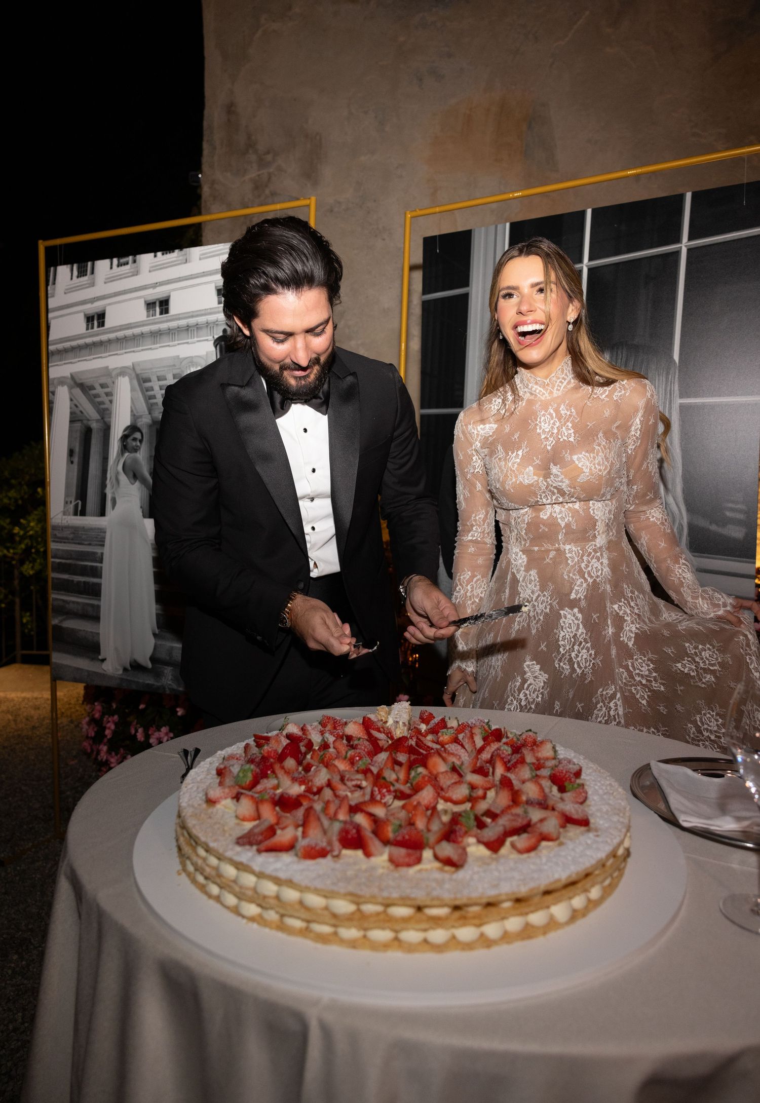 A couple in formal attire laughs while sharing a large strawberry tart dessert at an elegant dinner table.