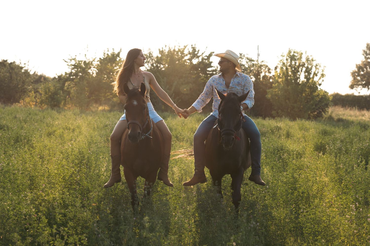 Two riders hold hands while horseback riding through a golden summer field at sunset.