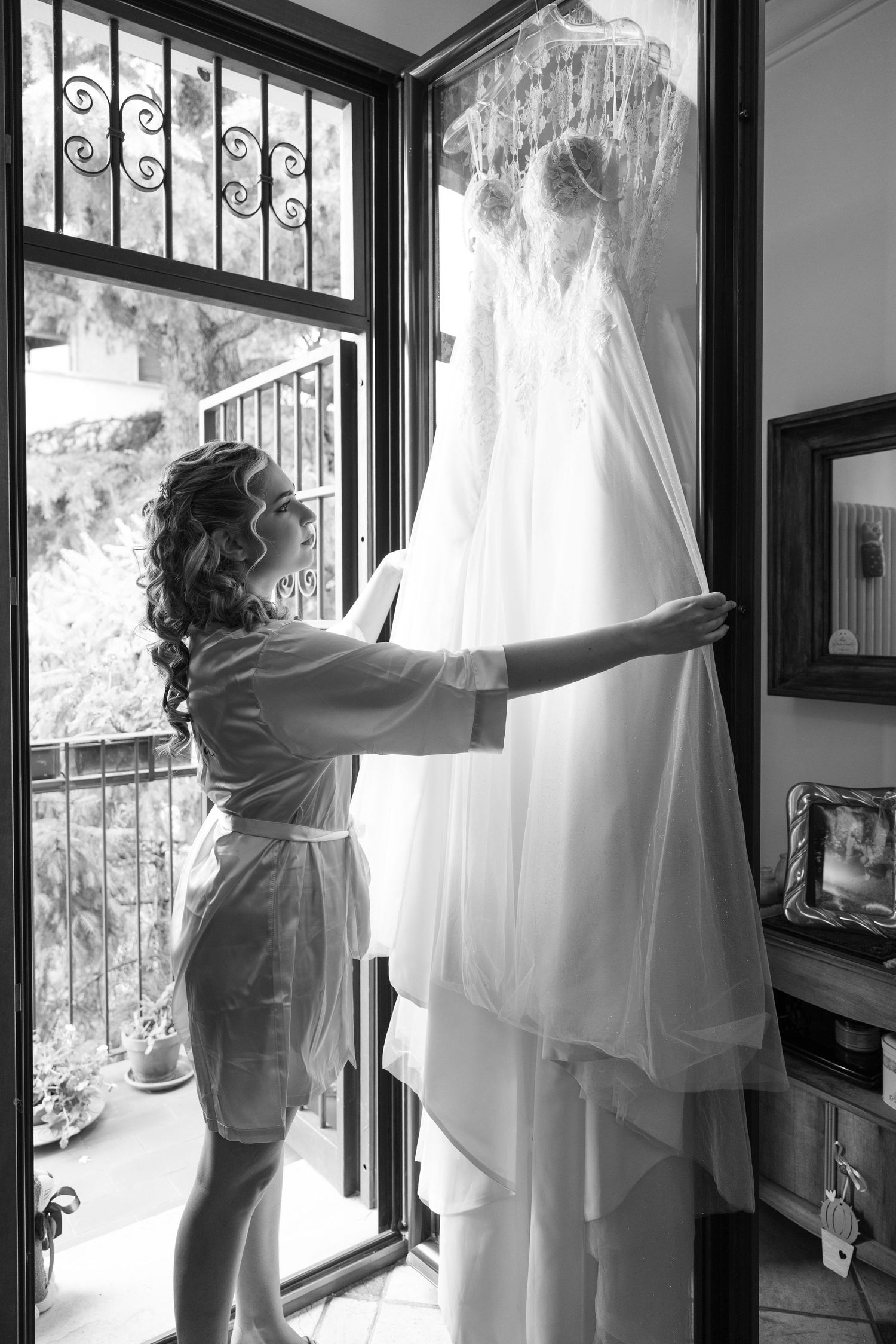 A black and white photo showing a wedding dress hanging by a window while someone adjusts it in morning light.