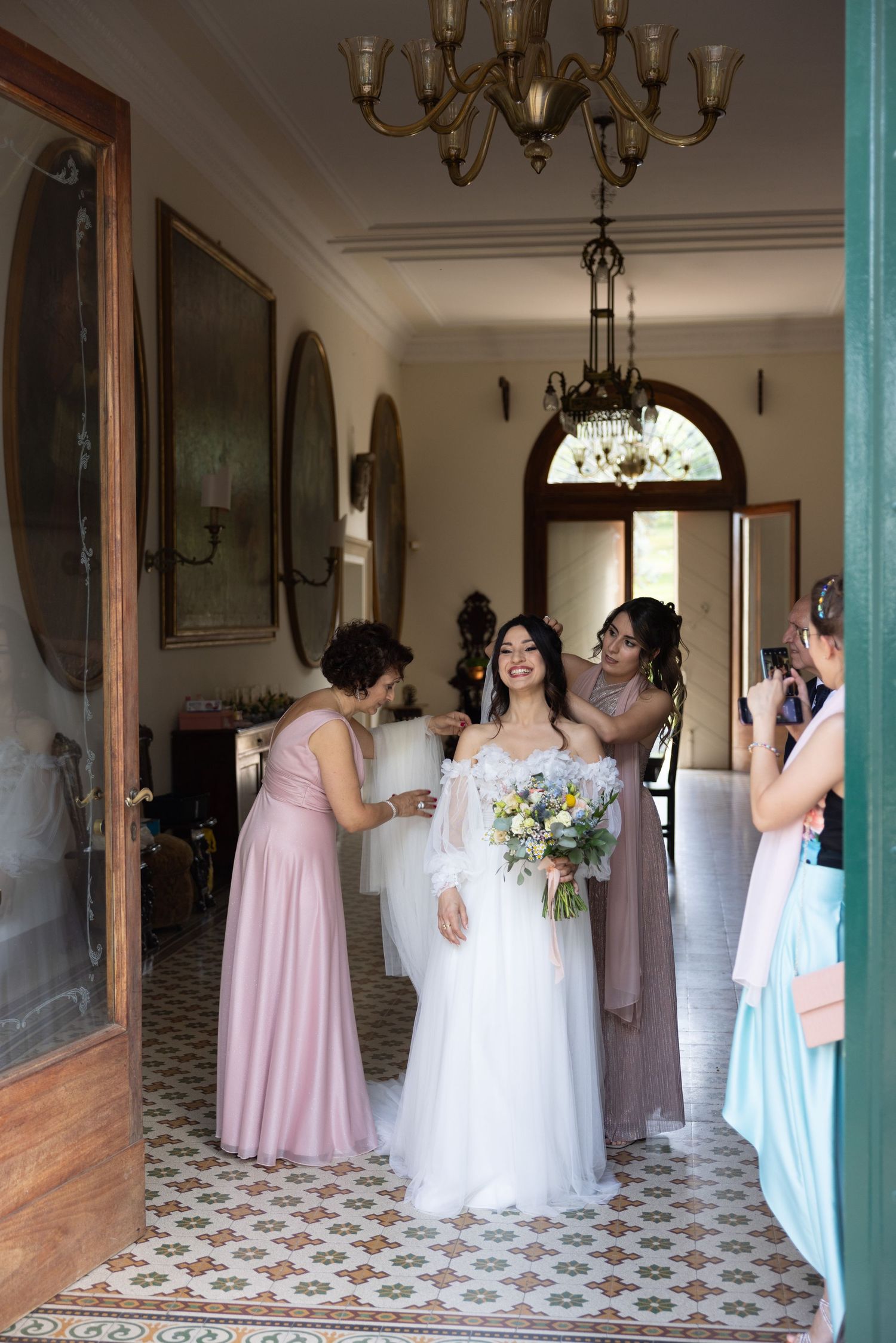 A wedding party gathers in an elegant room with patterned floors and chandelier.