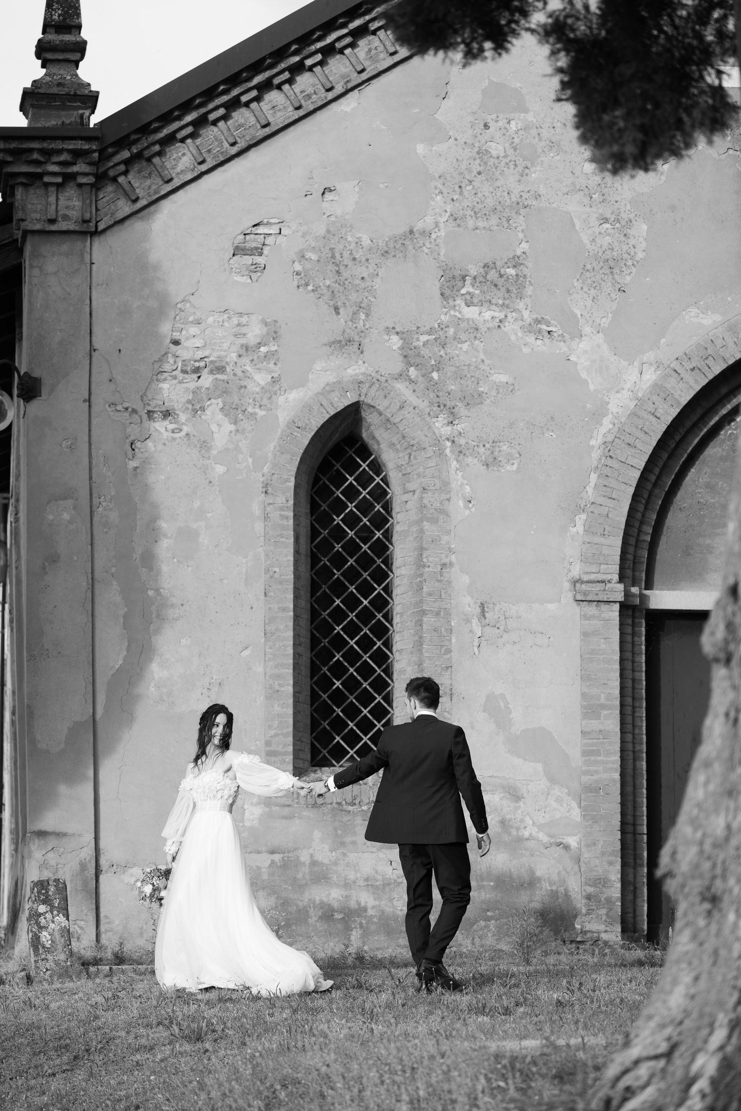 Black and white photo of a couple walking near an old church with arched windows and weathered walls.