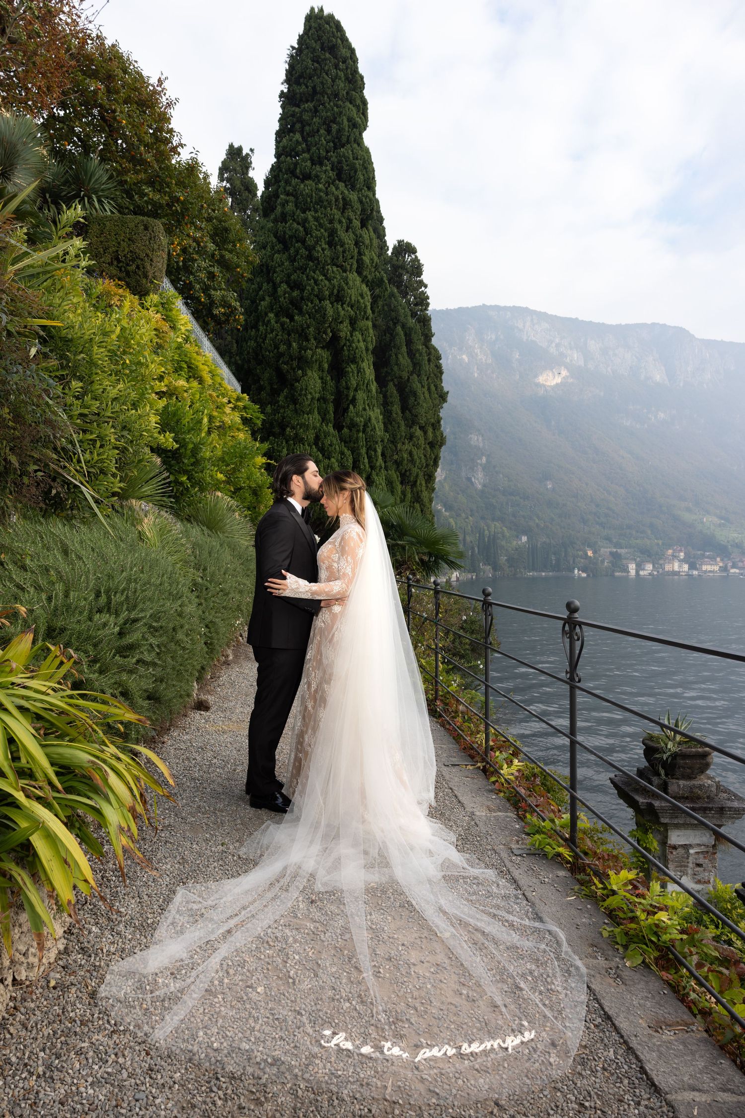 A romantic wedding couple poses on a scenic terrace overlooking Lake Como in Italy with cypress trees in the background.