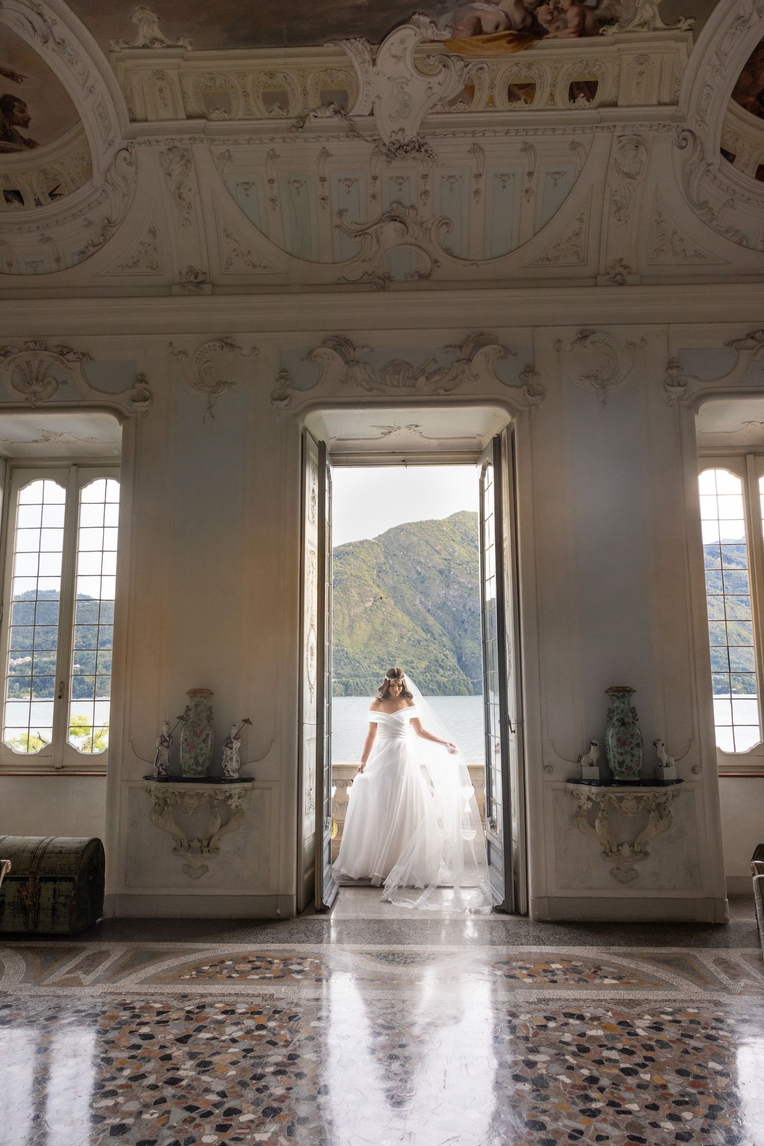 Wedding dress in elegant white room with painted ceiling and patterned tile floor overlooking lake.