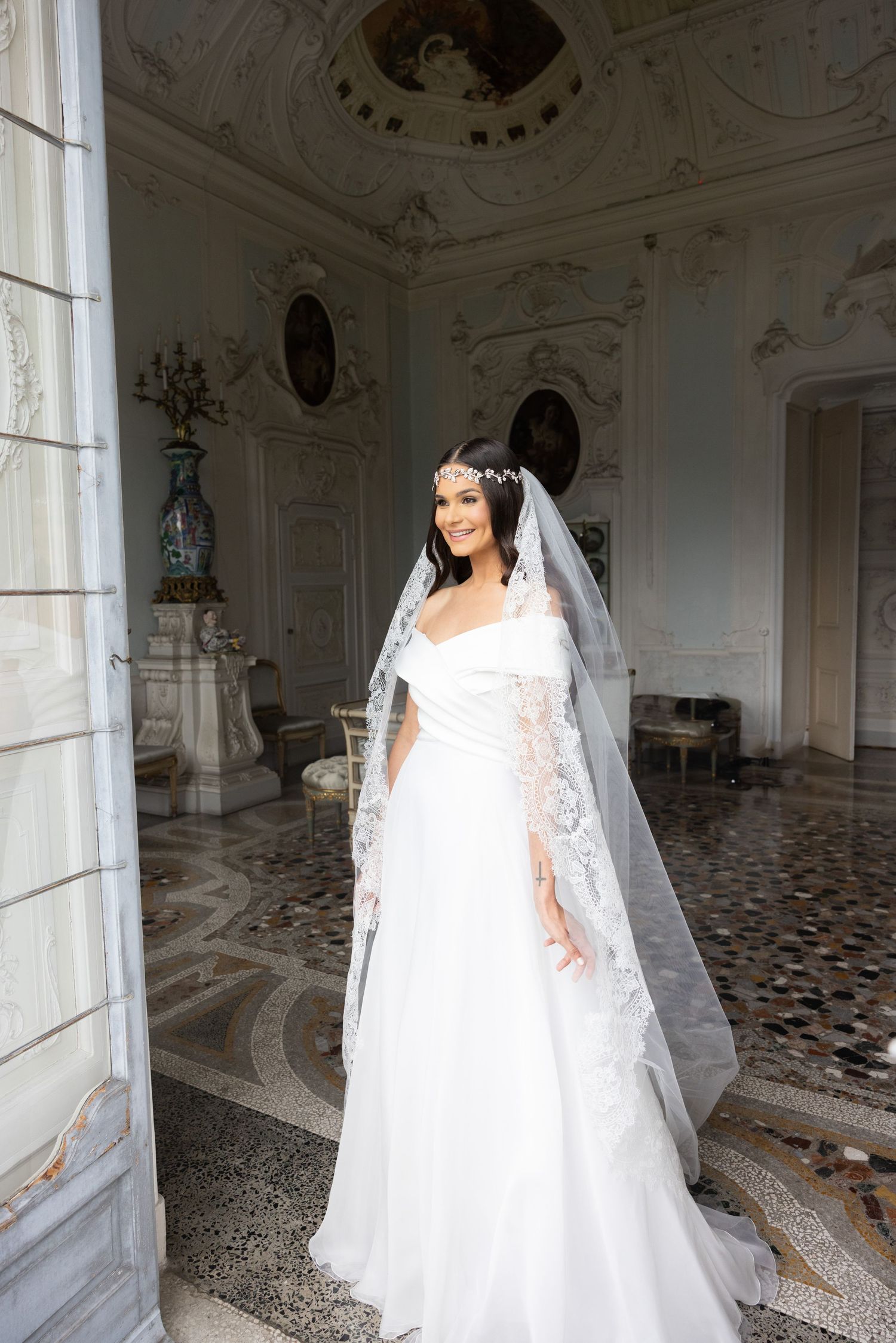 A bride wearing a flowing veil poses in a classical European-style room with painted ceilings.