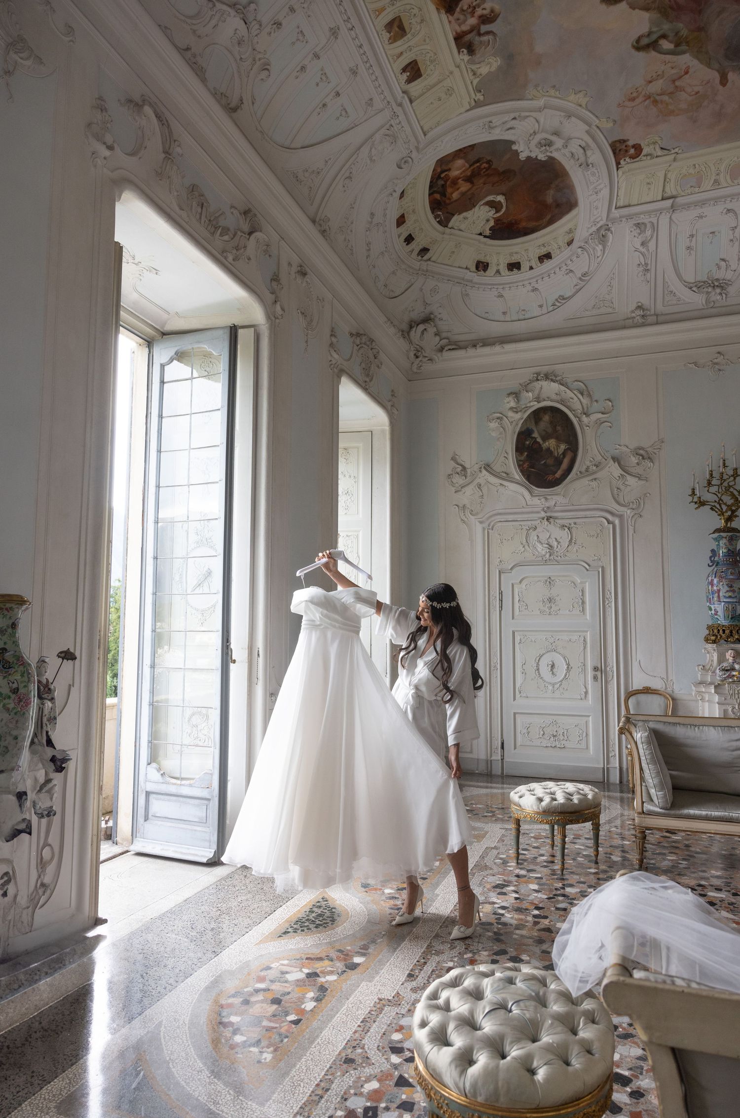 Wedding gown hanging in luxurious white room with ornate ceiling frescoes and tufted furniture.