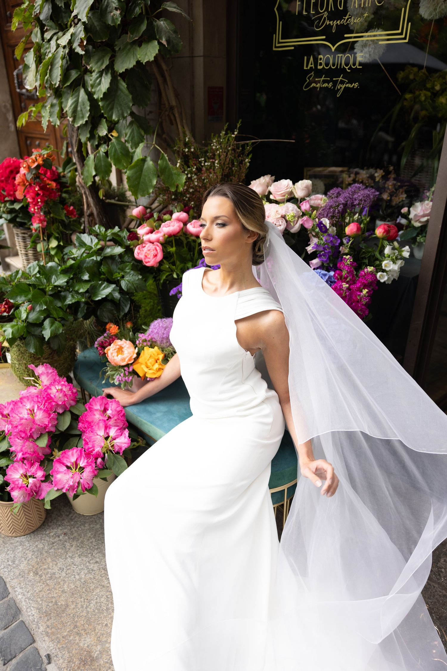 A bride in a white wedding dress poses elegantly among vibrant floral displays outside a boutique.