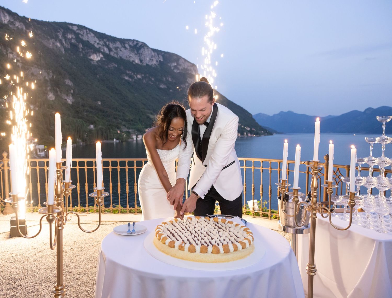 A couple cuts their wedding cake on an elegant terrace overlooking a scenic mountain lake at dusk with sparklers glowing.