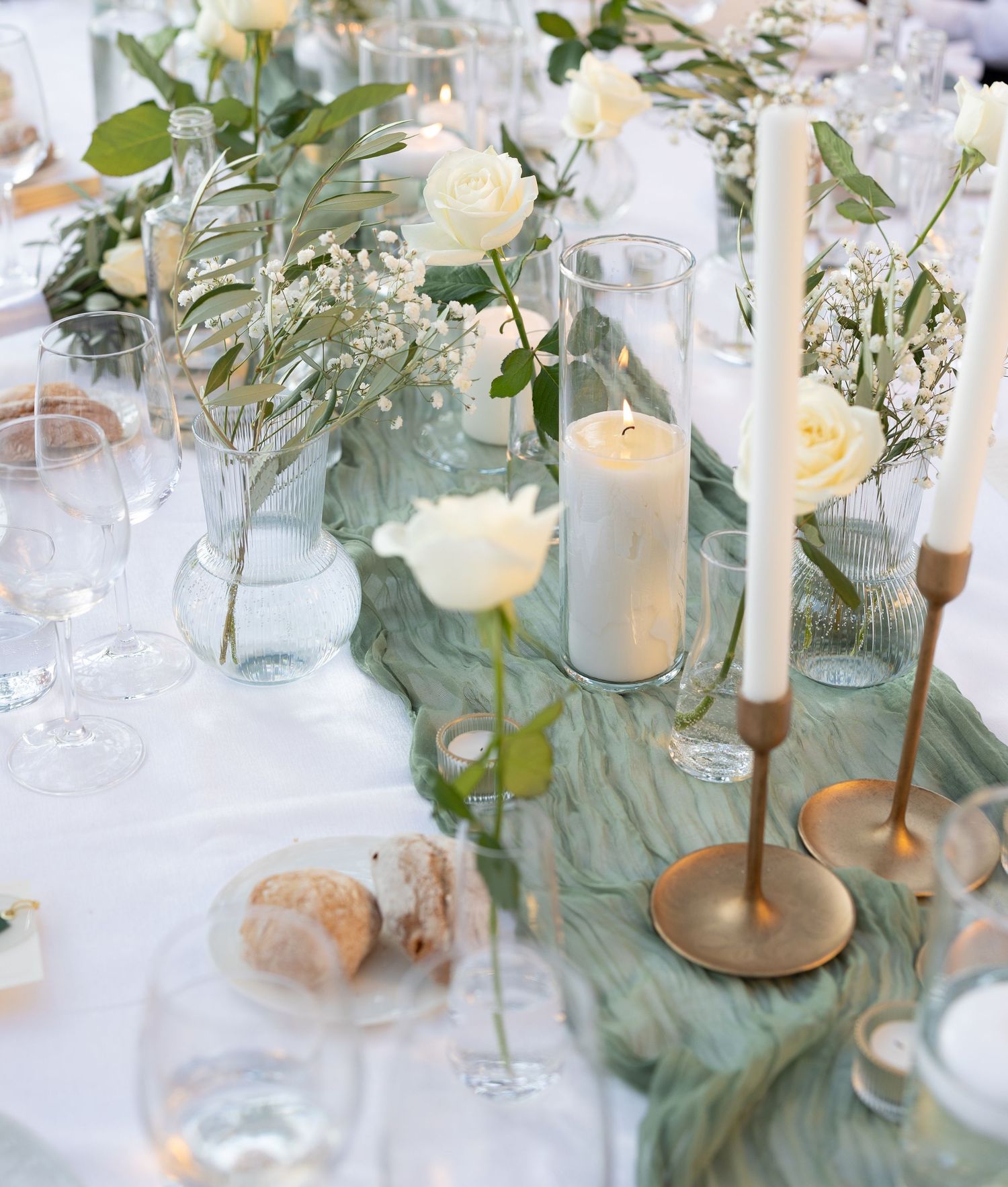 Elegant wedding table decor features white roses, eucalyptus leaves, and glowing candles in glass holders on a white tablecloth.