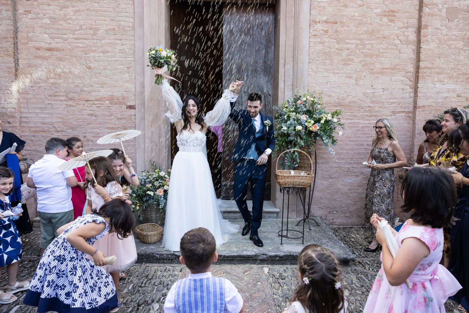 Outdoor wedding reception scene with guests mingling near a historic brick wall.