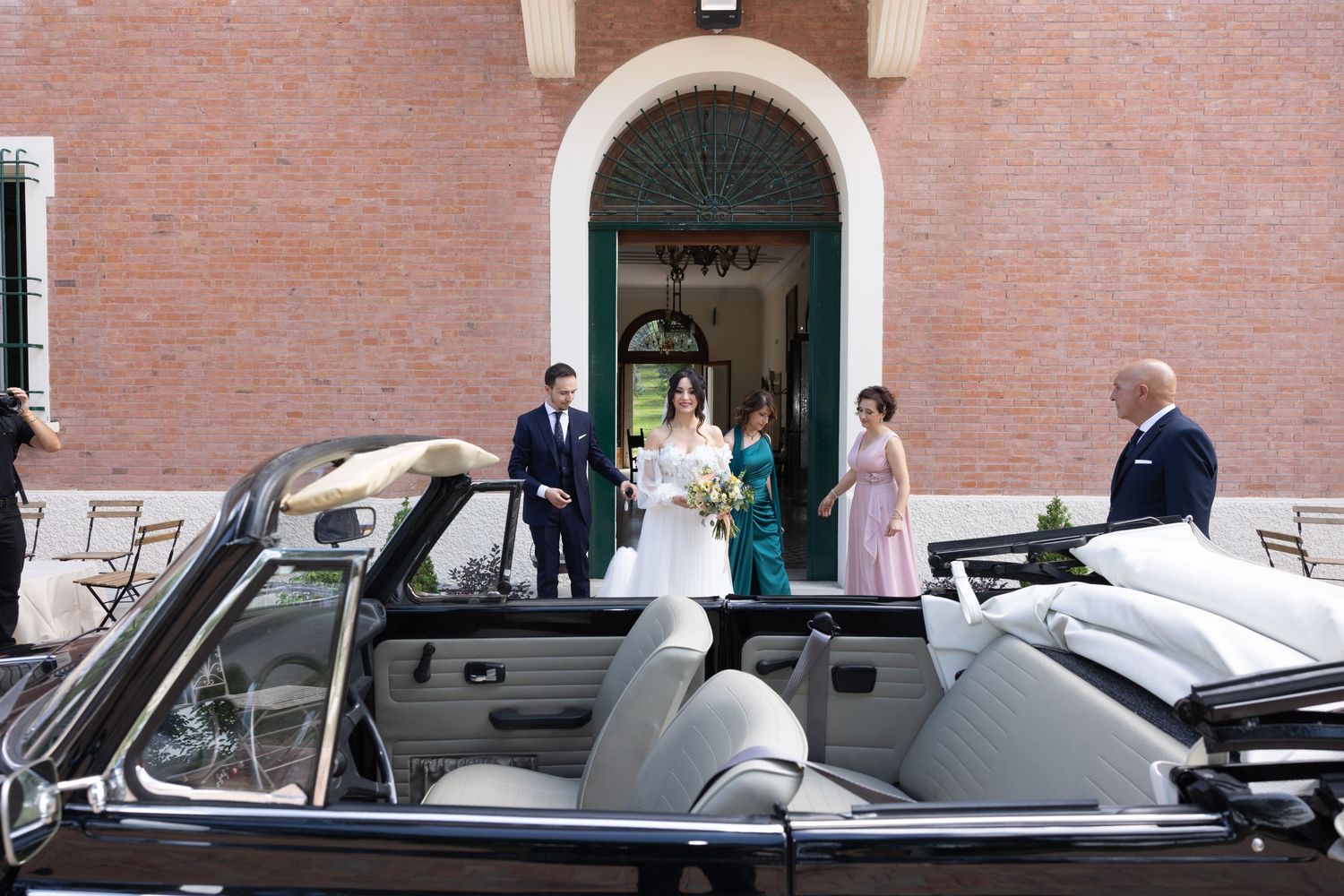 A vintage car awaits outside a brick building during a wedding celebration.