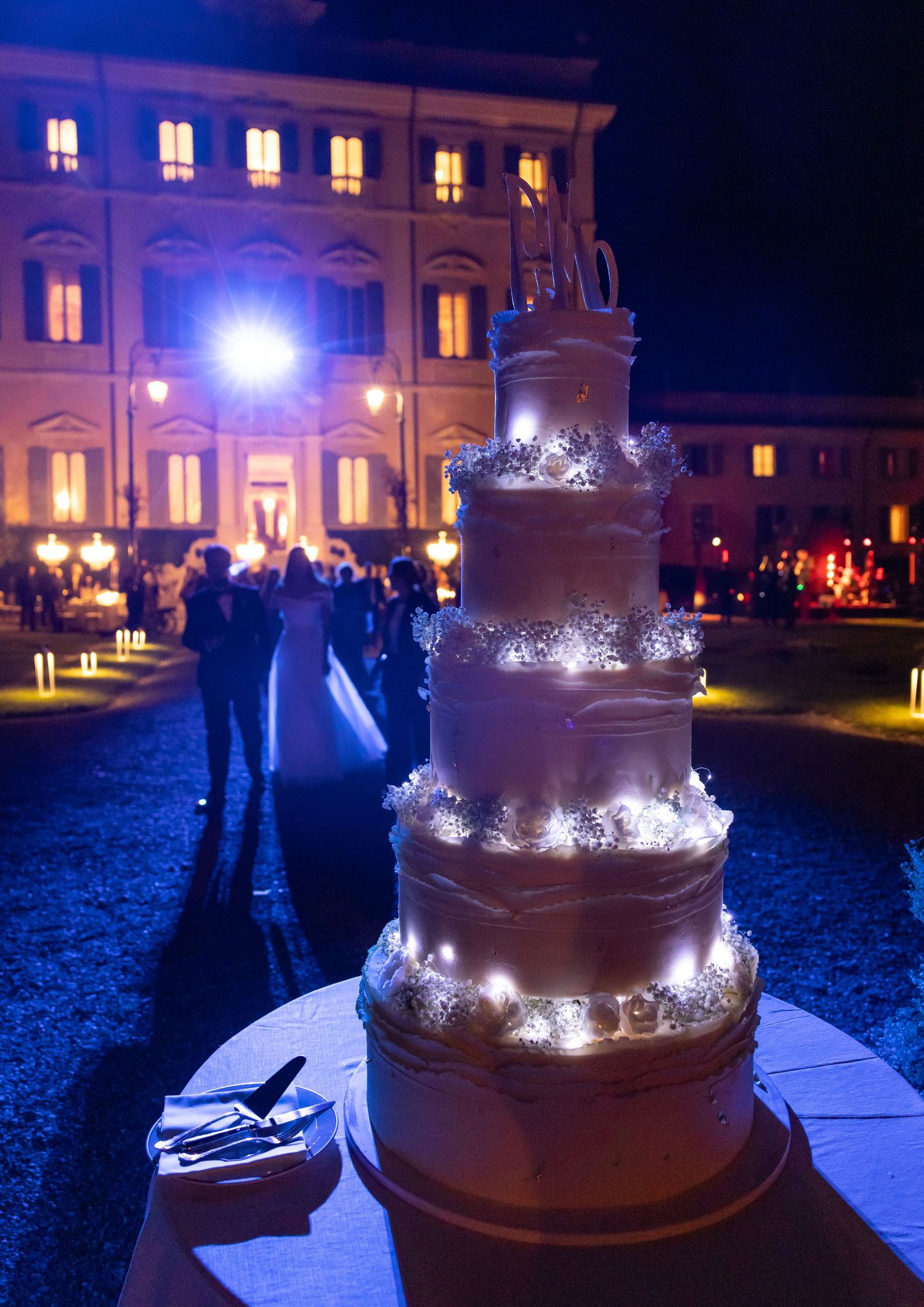 Three-tiered chocolate wedding cake with sparkling lights displayed outdoors at night.