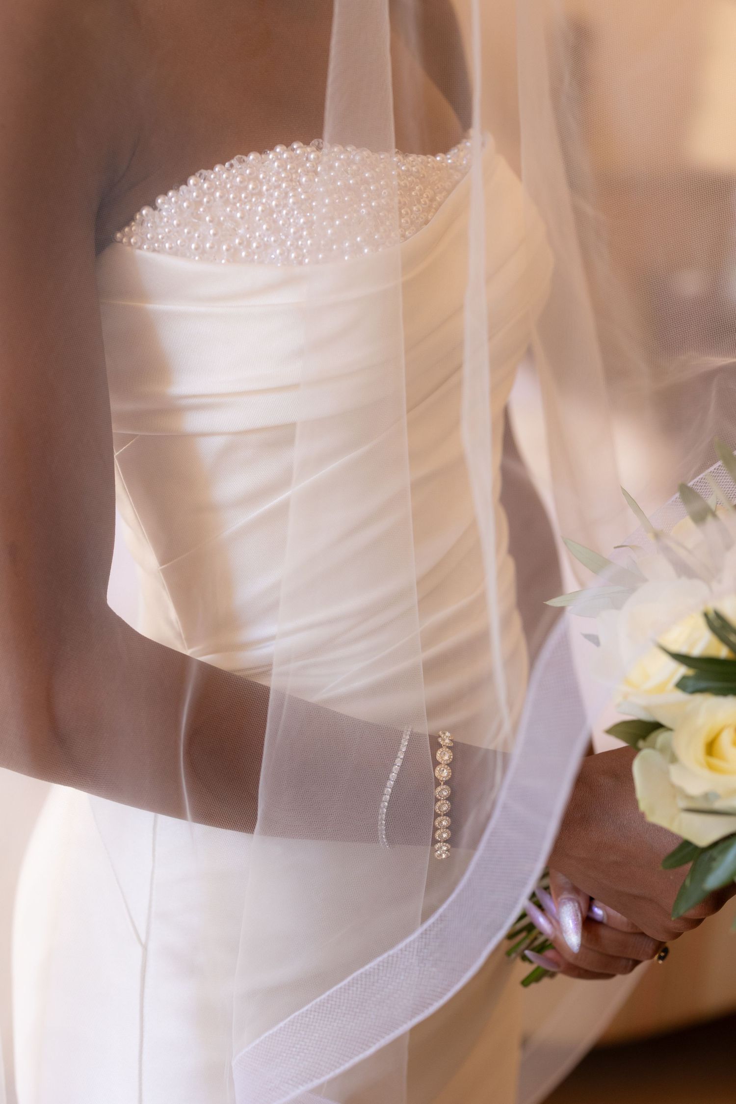 Close-up details of a white wedding dress showing delicate beading and ruched fabric texture in soft lighting.