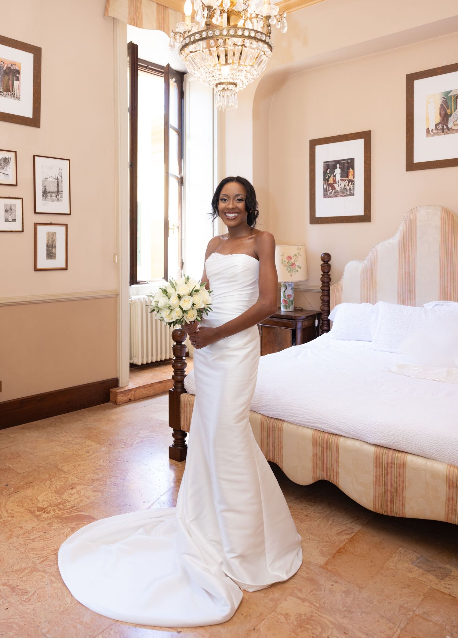 A bride in a flowing white strapless wedding gown poses in an elegant hotel room with crystal chandeliers and framed artwork.