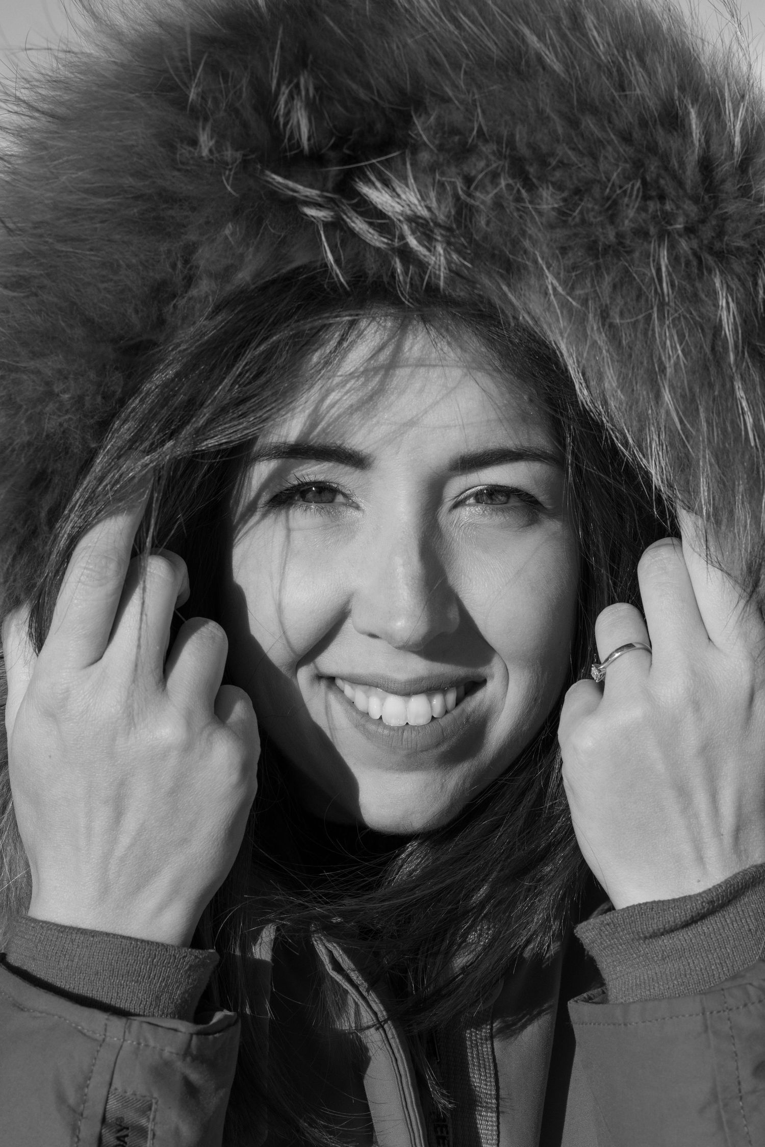 Black and white portrait of someone smiling while adjusting their furry winter hood.