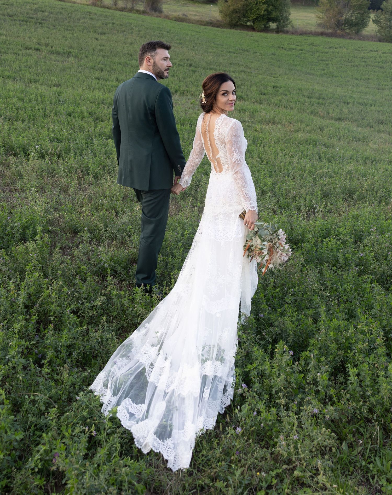 Wedding couple walking through a lush green field with flowing white lace dress trailing behind.