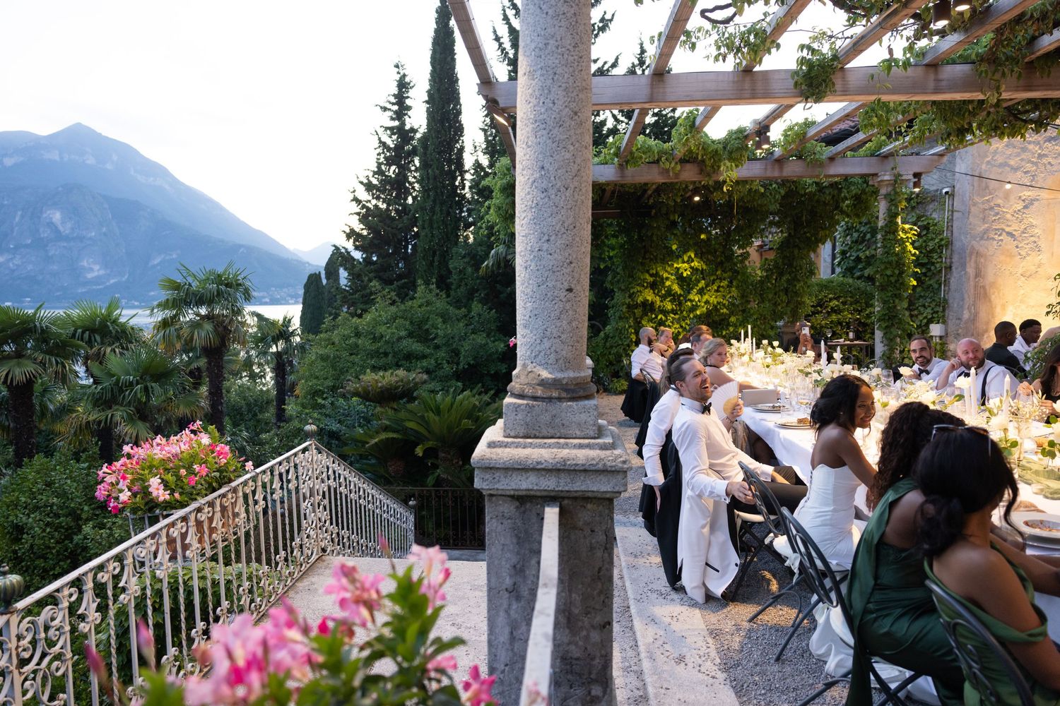 Wedding guests gather on a scenic mountain terrace overlooking a lake during an outdoor celebration at sunset.
