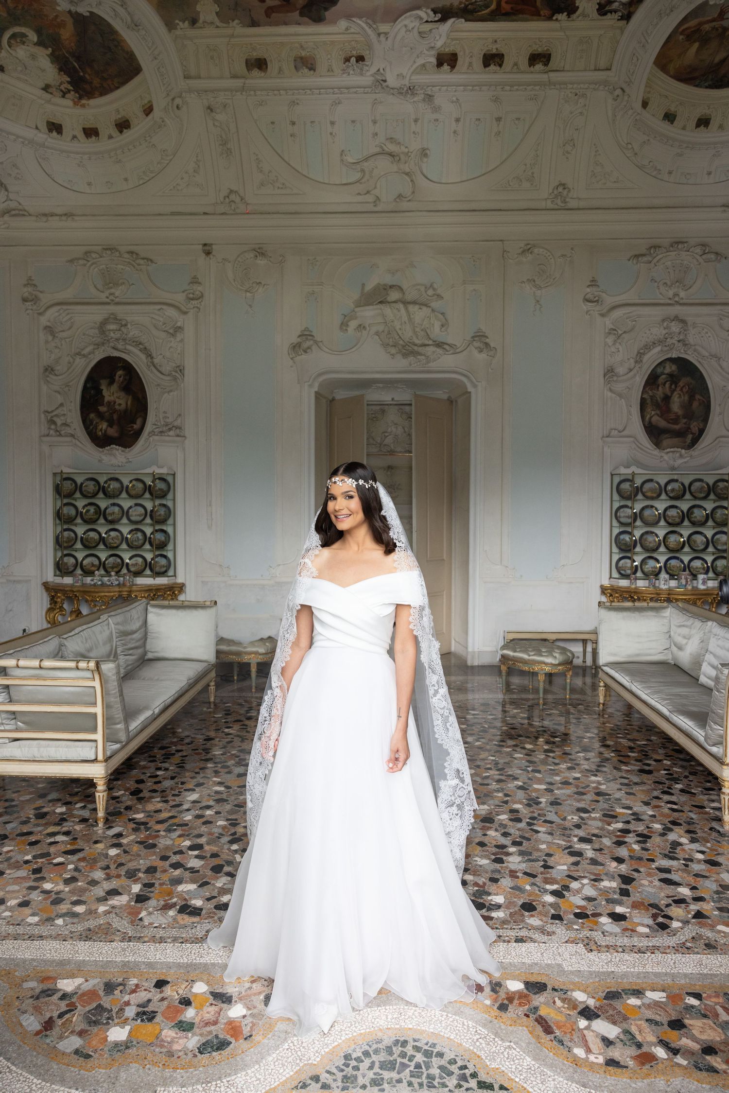 A bride stands in an elegant white gown against the backdrop of a historic room with ornate architectural details.